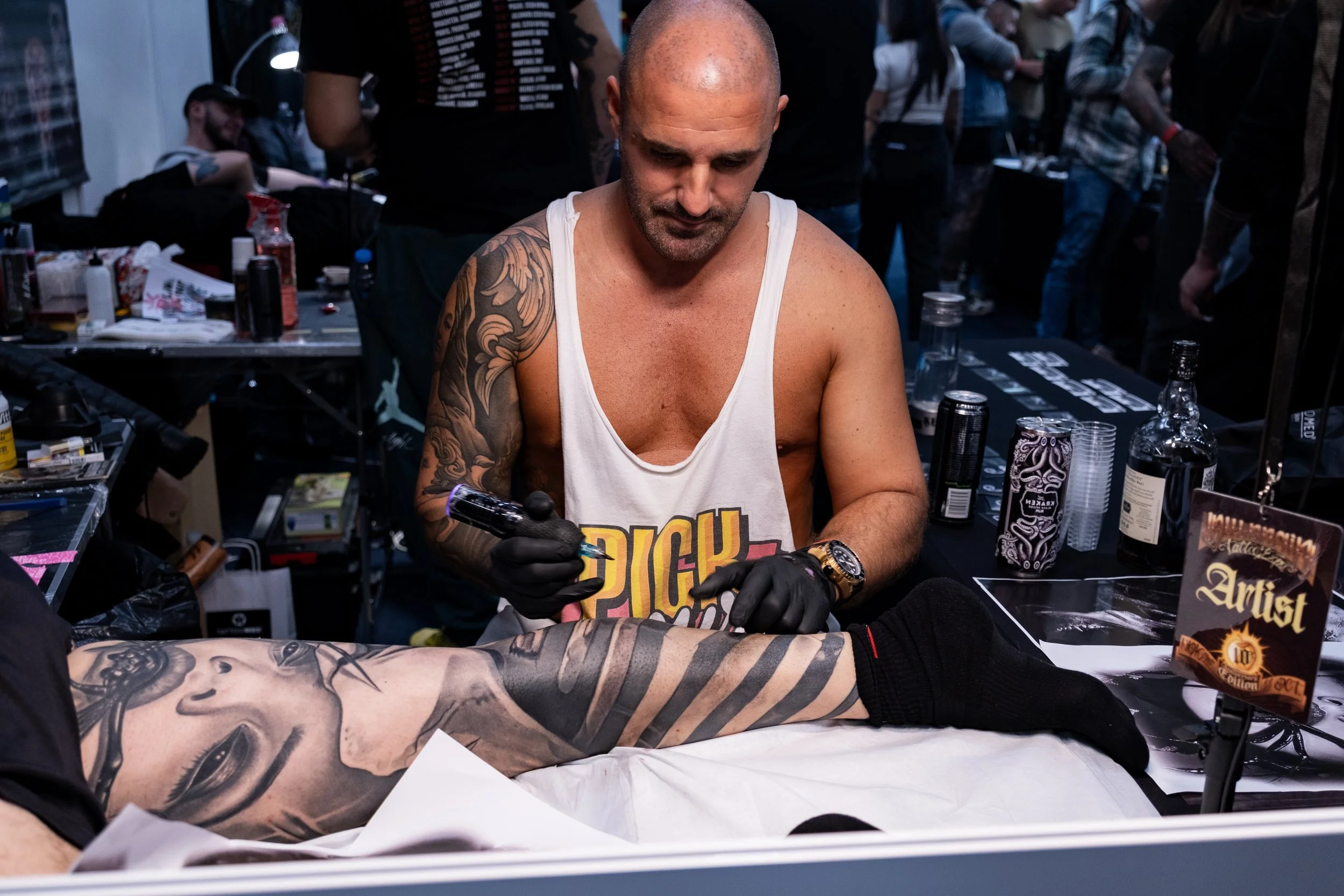 A tattoo artist working on a person's leg at a tattoo convention, with various supplies on the table and other attendees in the background.