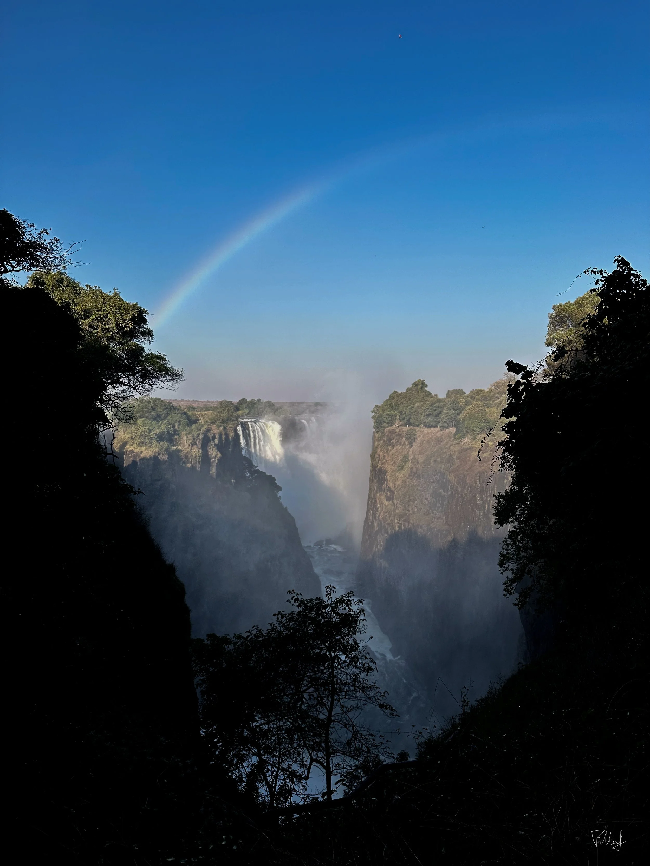View of a waterfall with mist and a rainbow in the sky, surrounded by trees and cliffs.