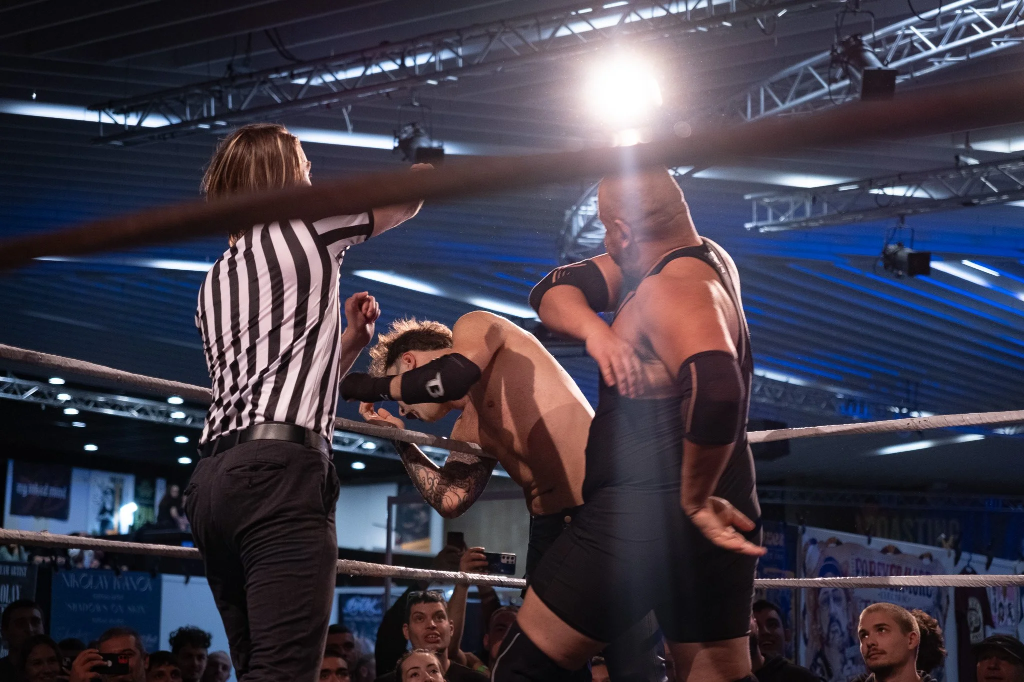 Two wrestlers compete in a match while a referee watches and an audience observes in the background.