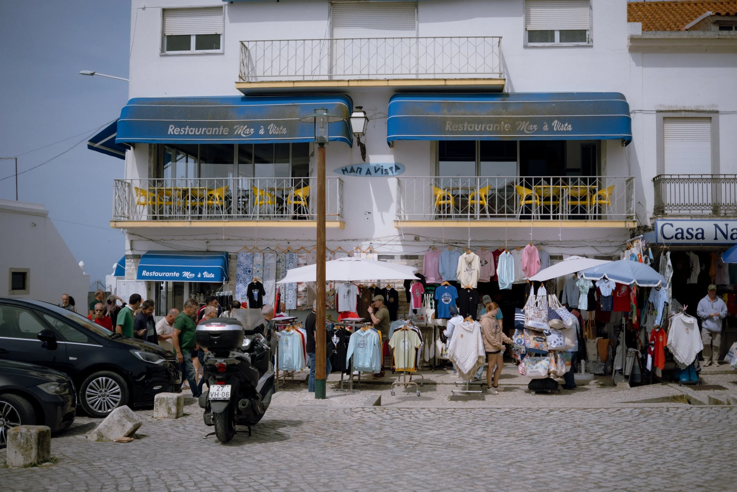 Street view of a two-story white building with outdoors market stalls selling clothes in front. People browse, cars and a motorcycle are parked nearby. Blue awnings display the name 'Restaurante Mar à Vista'.