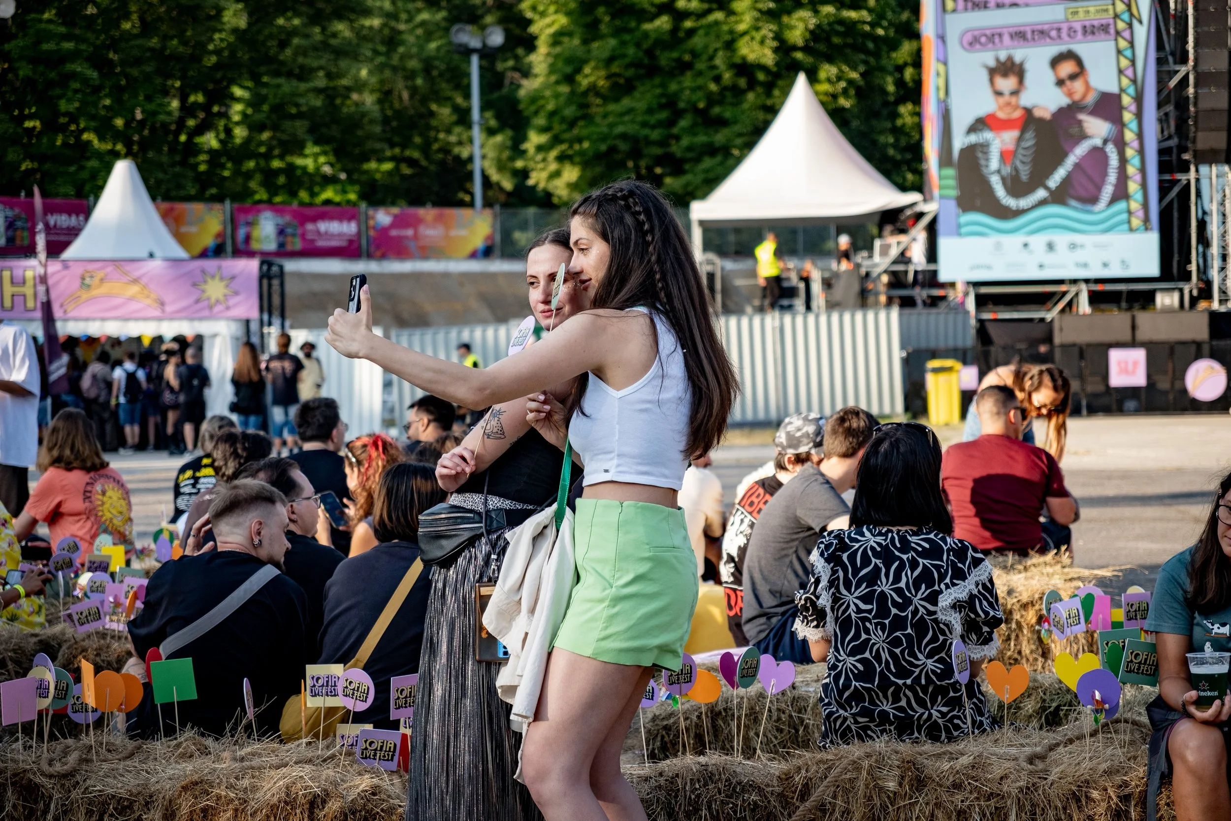 Two young women taking a selfie at an outdoor music festival with a stage and crowd in the background, decorated with colorful signs and hay bales
