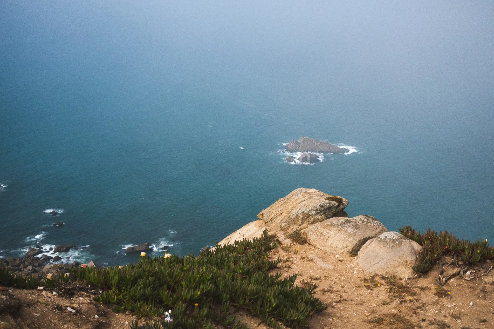 A view from a cliff overlooking a misty ocean with rocks and a small boat in the distance.