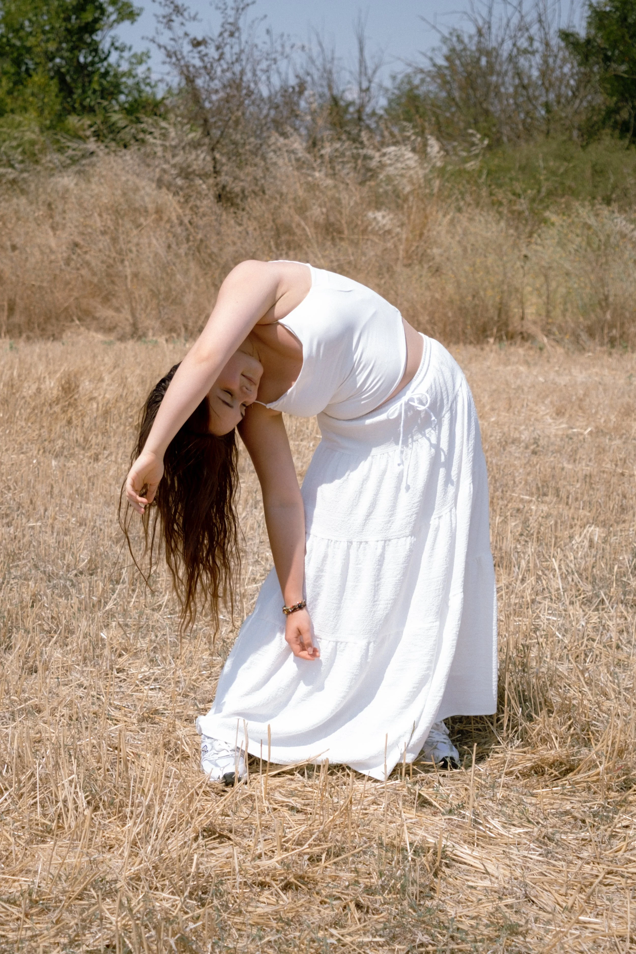 Portrait photography. Woman with long brown hair wearing a white tank top and long white skirt, bending backward in a field of dry grass with trees in the background.