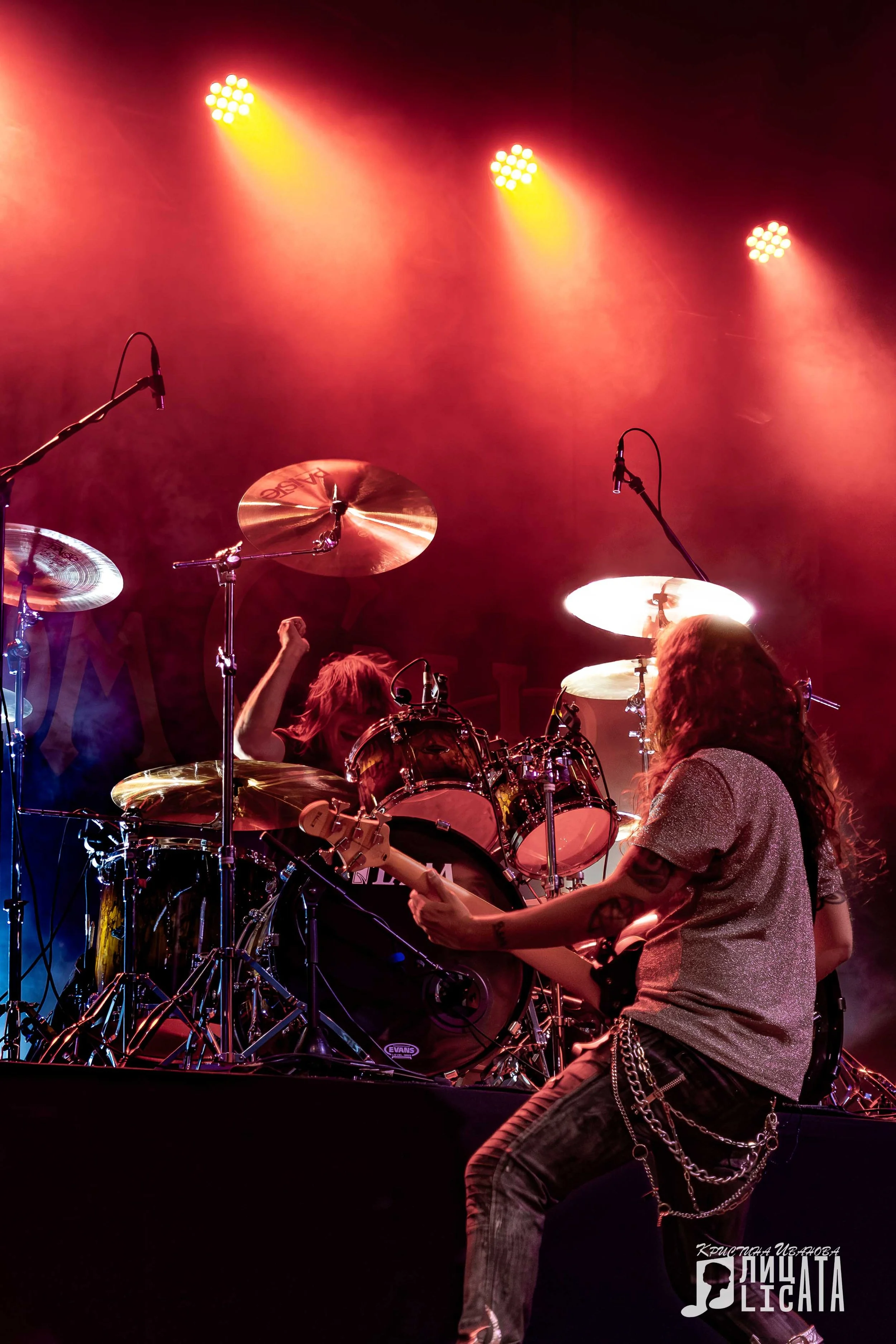 A rock band performing on stage with a drummer, guitarist, and bright red and yellow stage lighting.