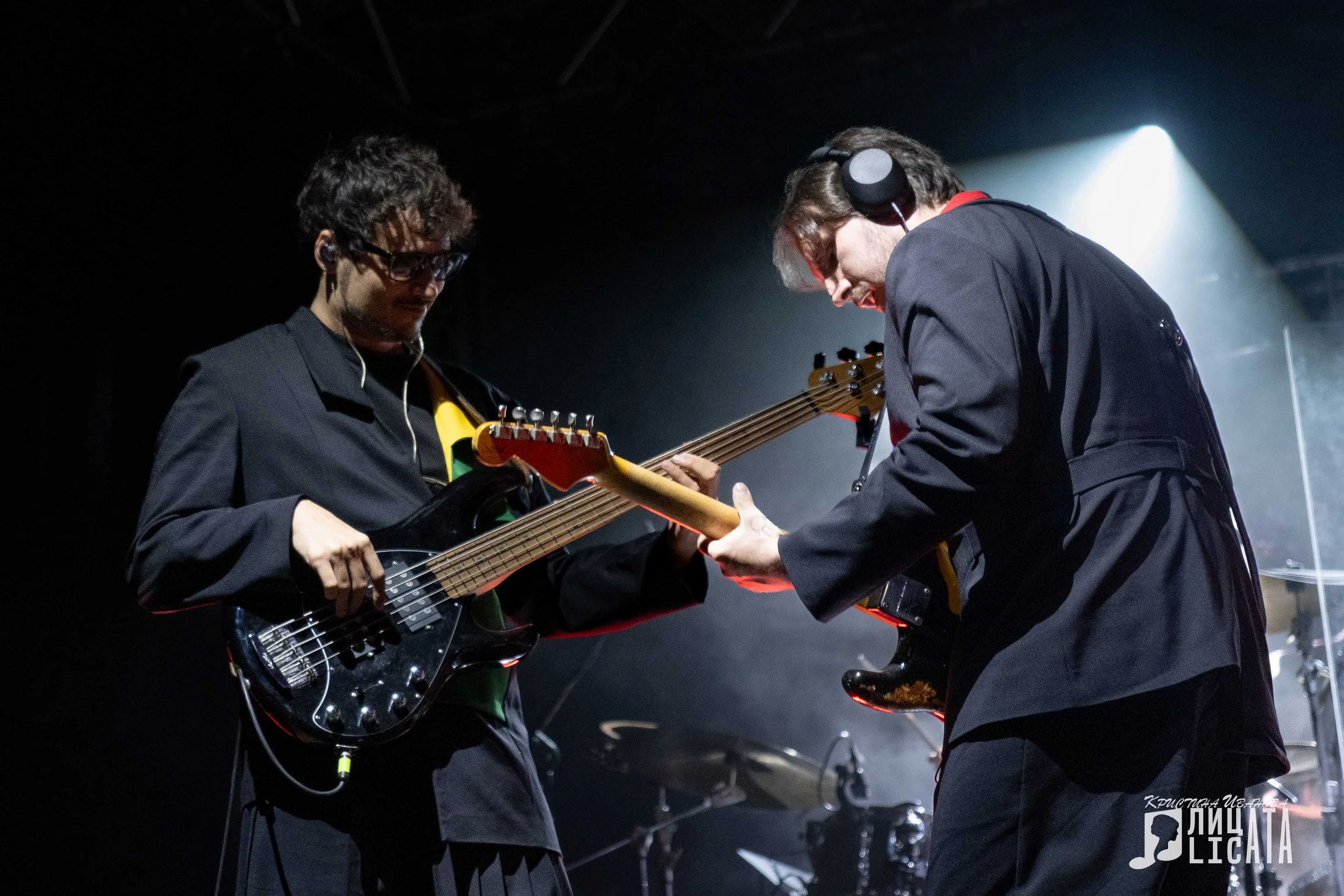 Two musicians playing guitars on a dimly lit stage, one with glasses and curly hair, and the other with headphones and glasses, both focused on their instruments.