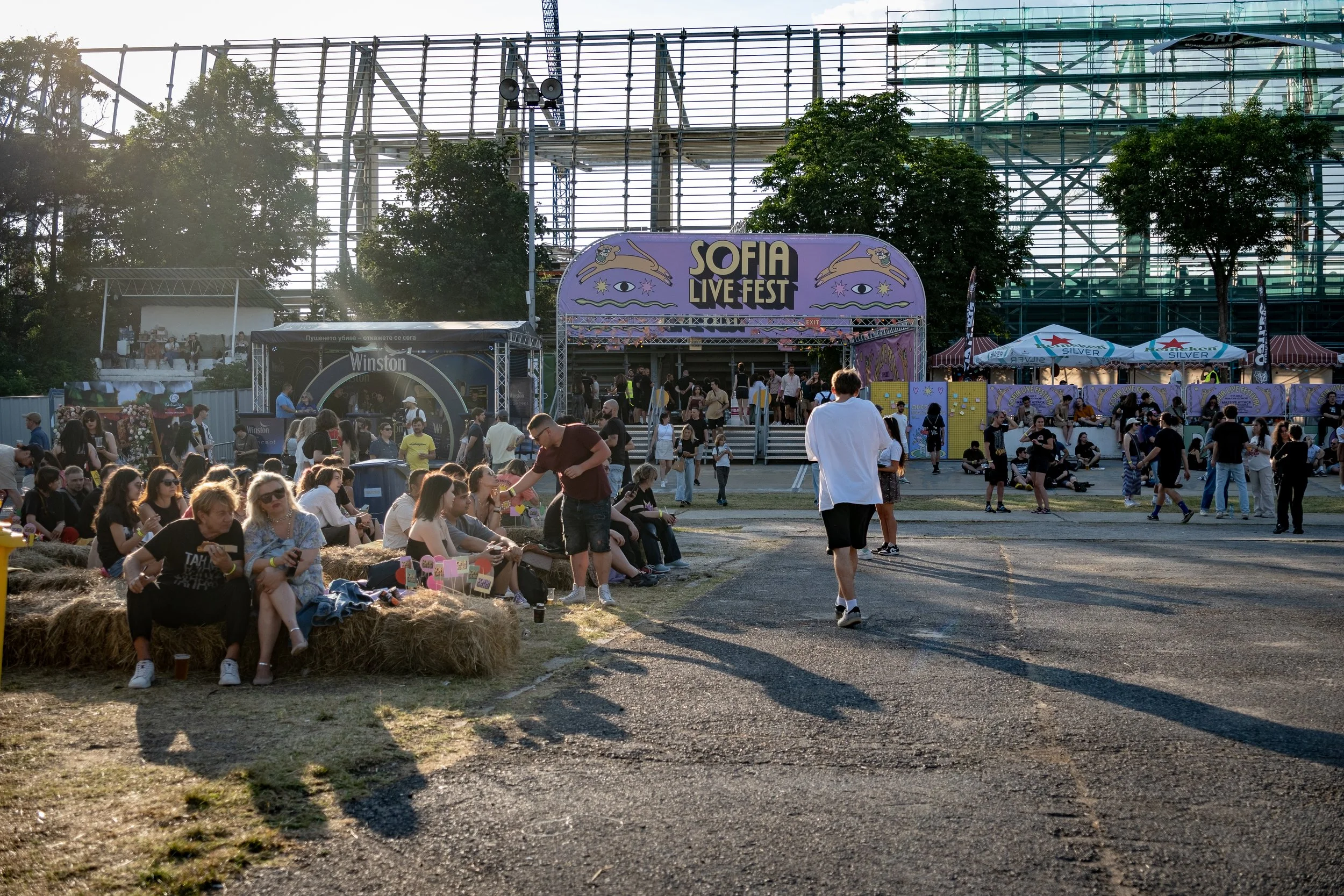 People attending Sofia Live Fest outdoor music event in the late afternoon. Some are sitting on hay bales, others walking or standing near stages and tents. The main stage has a decorated arch with the festival name, and there are trees and industria