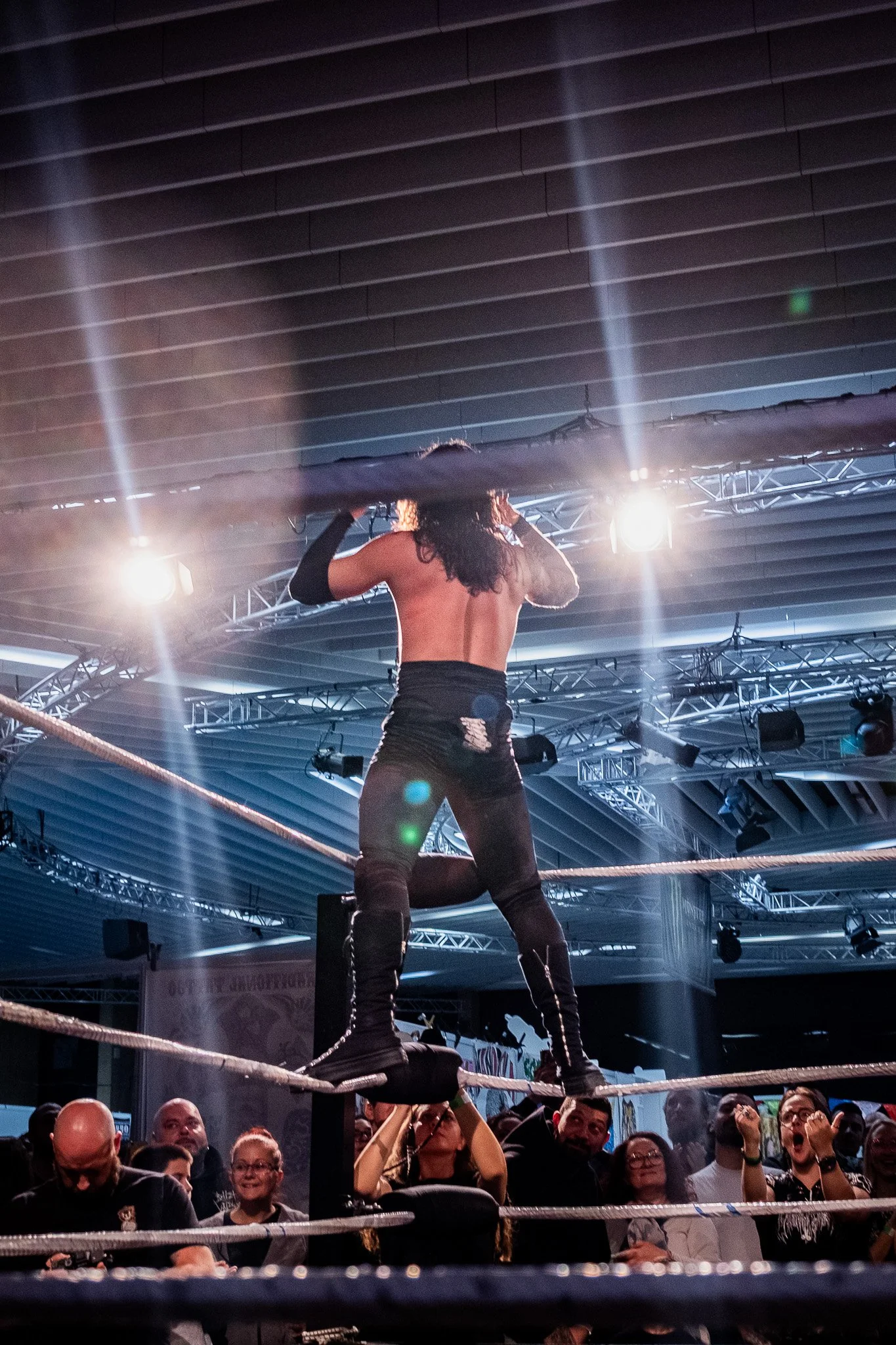A professional wrestling event with a muscular wrestler standing on the top turnbuckle in the ring, holding a black cloth over his shoulders, and an audience cheering in the background under dramatic lighting.