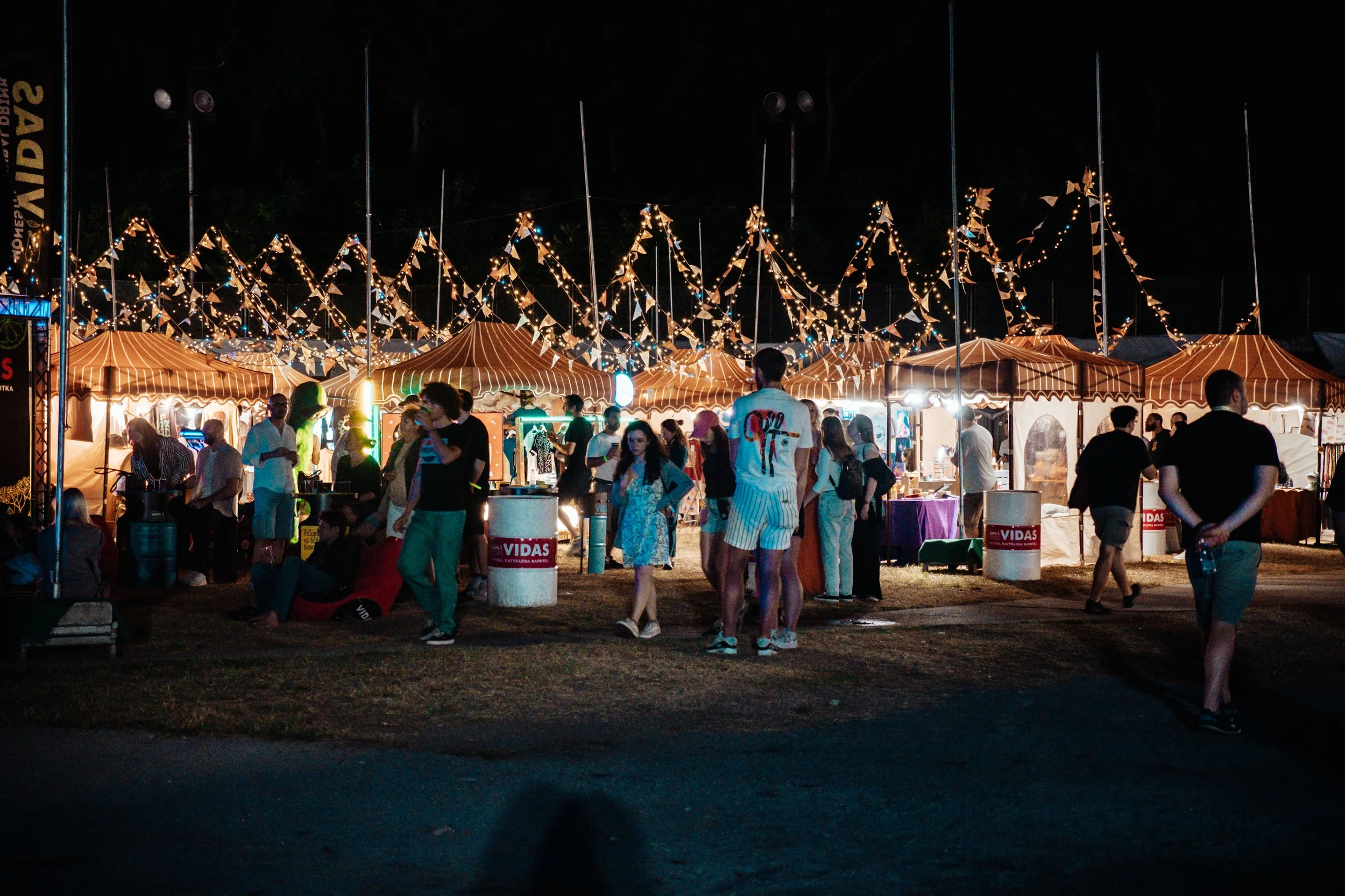 People gathered at night under string lights and tents at an outdoor festival or fair, with food stalls and booths.