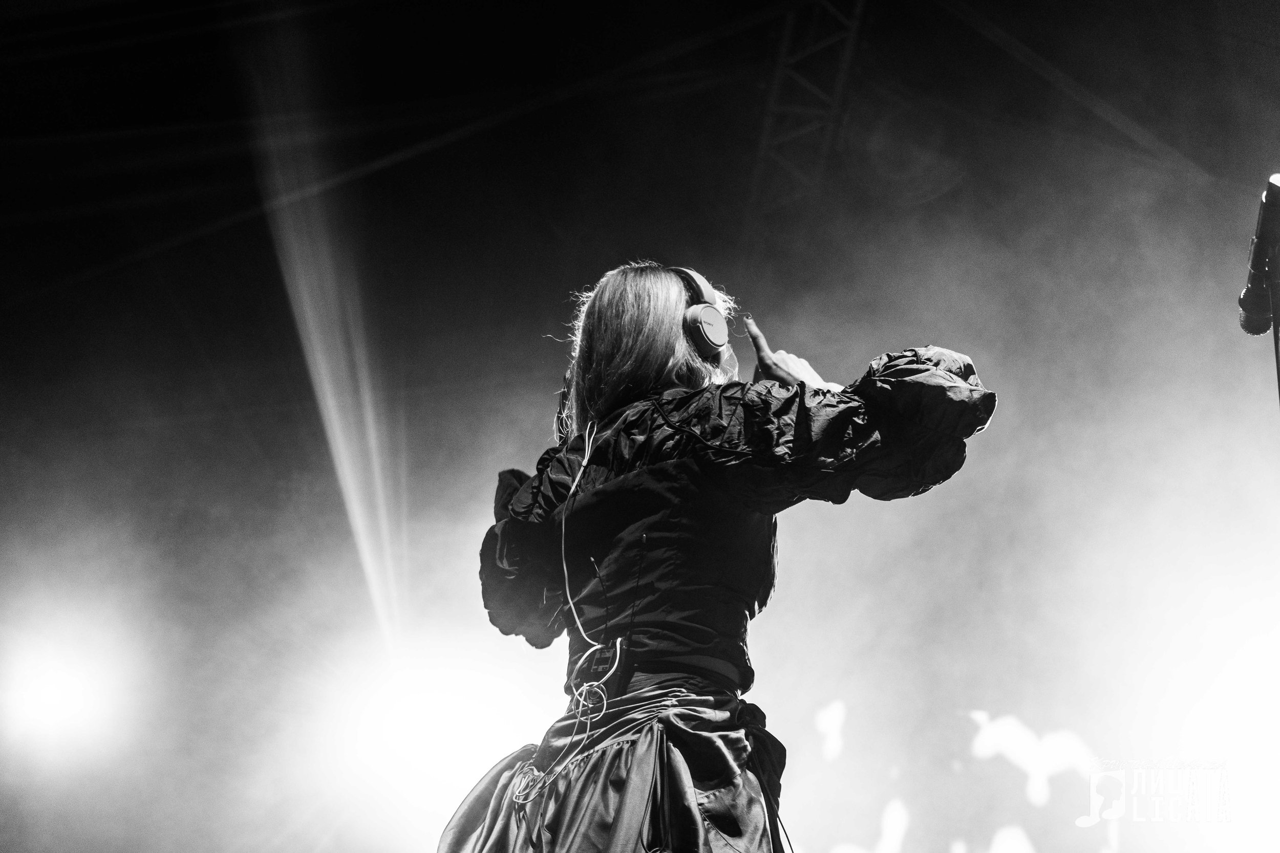 A woman with long hair wearing headphones, standing on stage with fog and bright lights, adjusting her headphones.