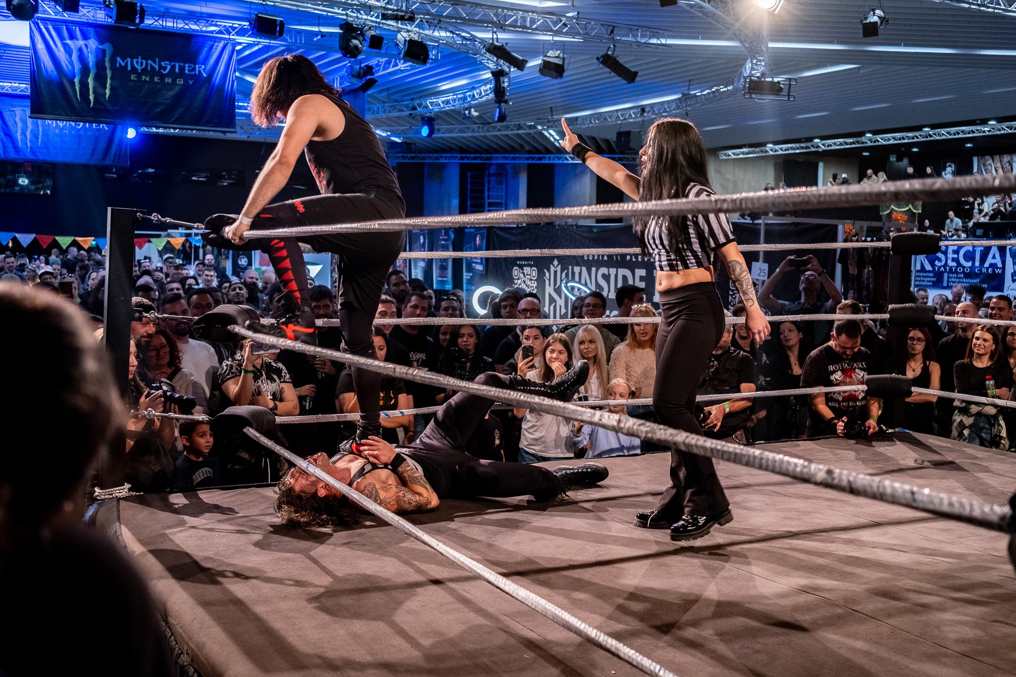 A professional wrestling match with one wrestler on the ground and another standing on her, while a referee signals in the ring. The crowd watches intently.