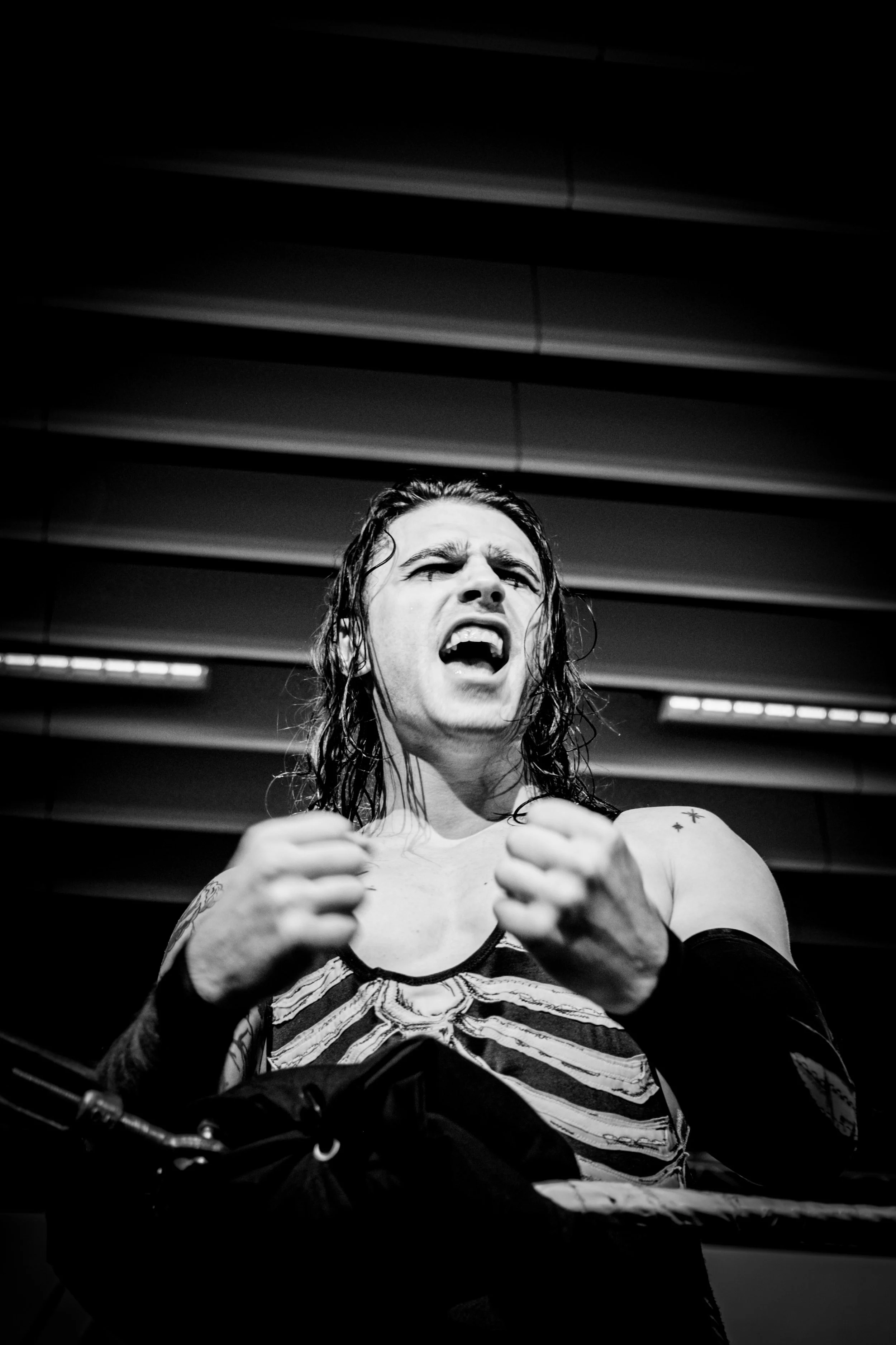 A black and white photo of a male wrestler in a fighting stance, with long dark hair, wearing a skeleton-themed outfit, inside a wrestling ring.
