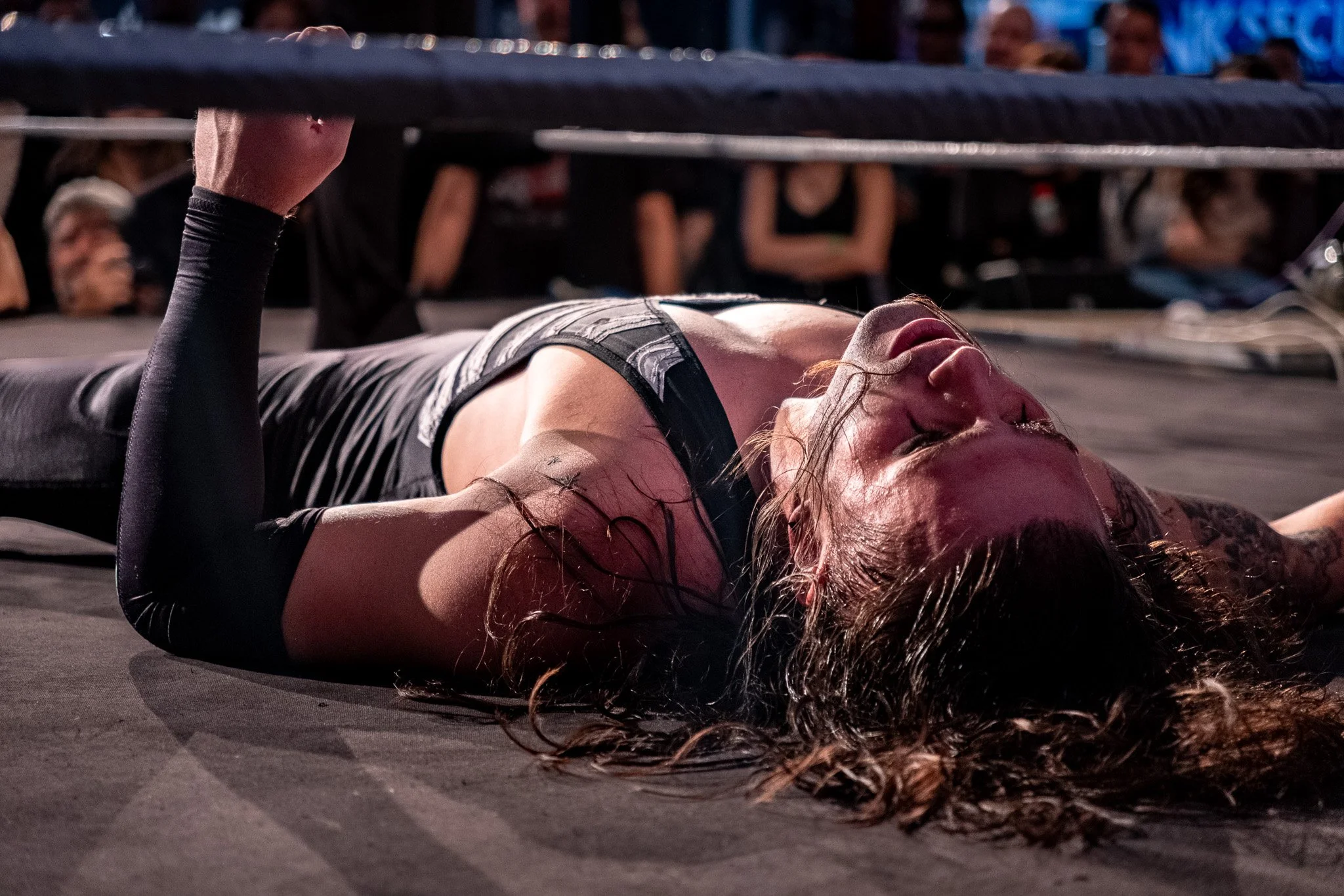 A female wrestler lying on the mat after a match, appearing exhausted, with her arm raised in victory, in a wrestling ring surrounded by spectators.