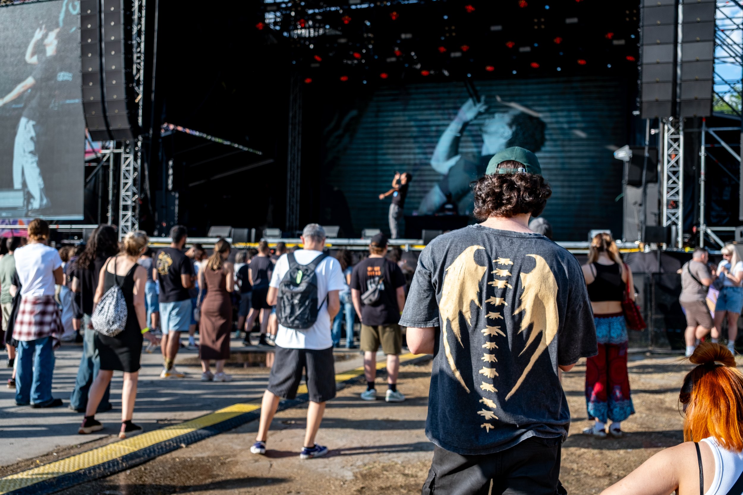 Crowd of people at an outdoor concert or festival, watching a performer on stage with a large screen behind them, during daylight.