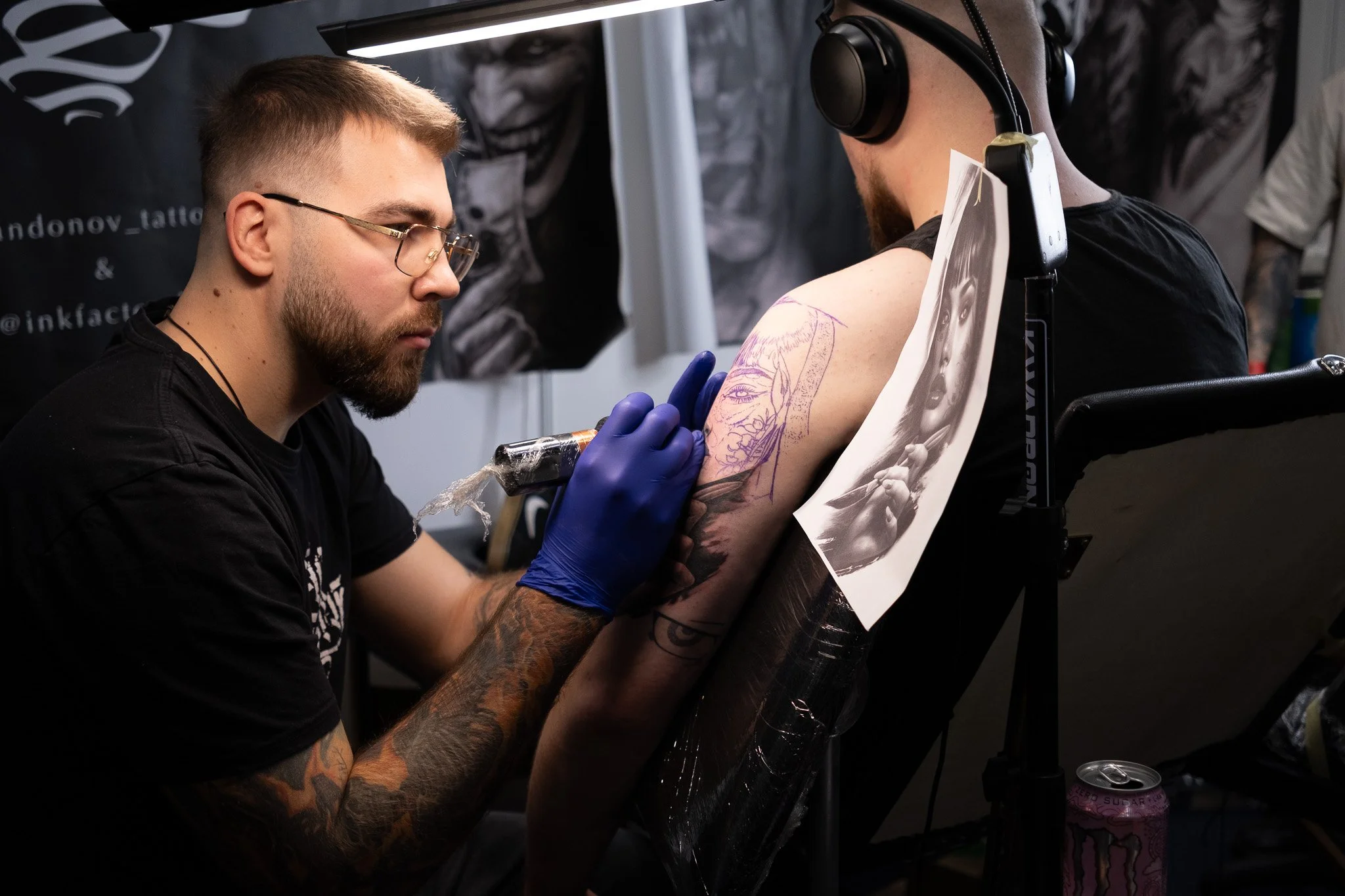 Tattoo artist creates a tattoo on a person's upper arm, with the process shown with reference images pinned to the chair.