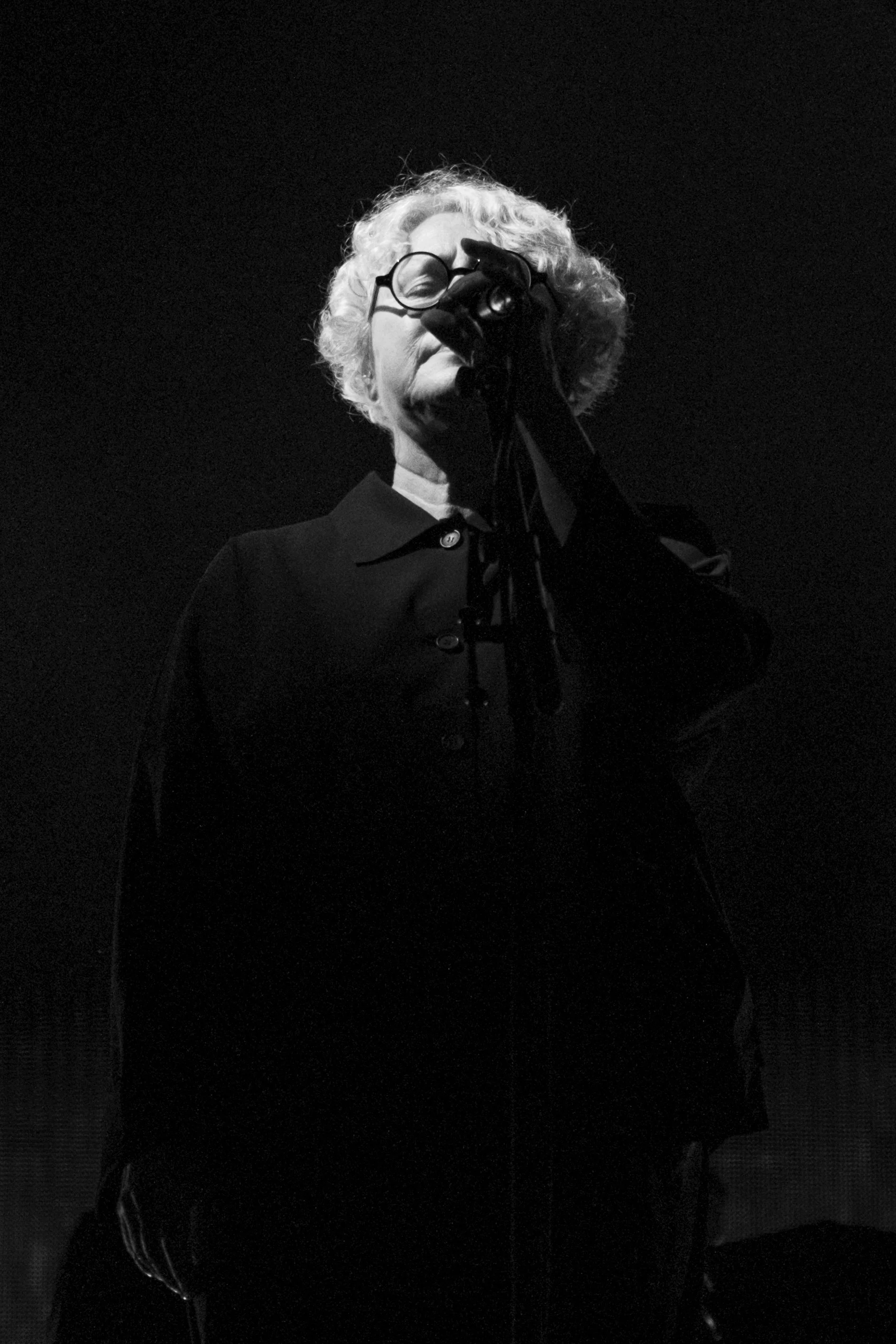 Concert Photographer captured Elizabeth Fraser singing with Massive Attack band at Sofia Live Festival. Black and white photo of an elderly woman with curly hair, wearing glasses and a dark outfit, singing into a microphone on a dark stage.