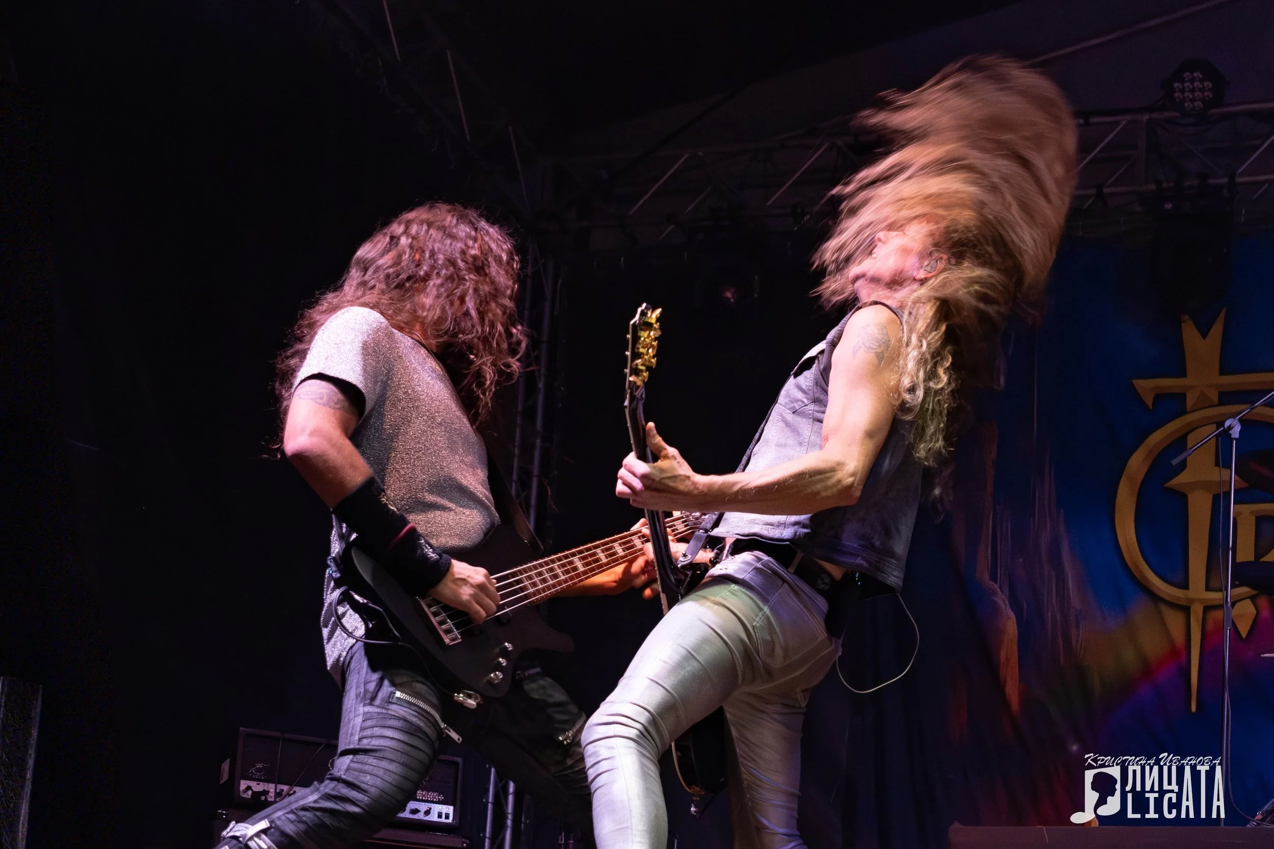 Two men playing electric guitars on stage during a concert, with dynamic hair movement and dramatic lighting.