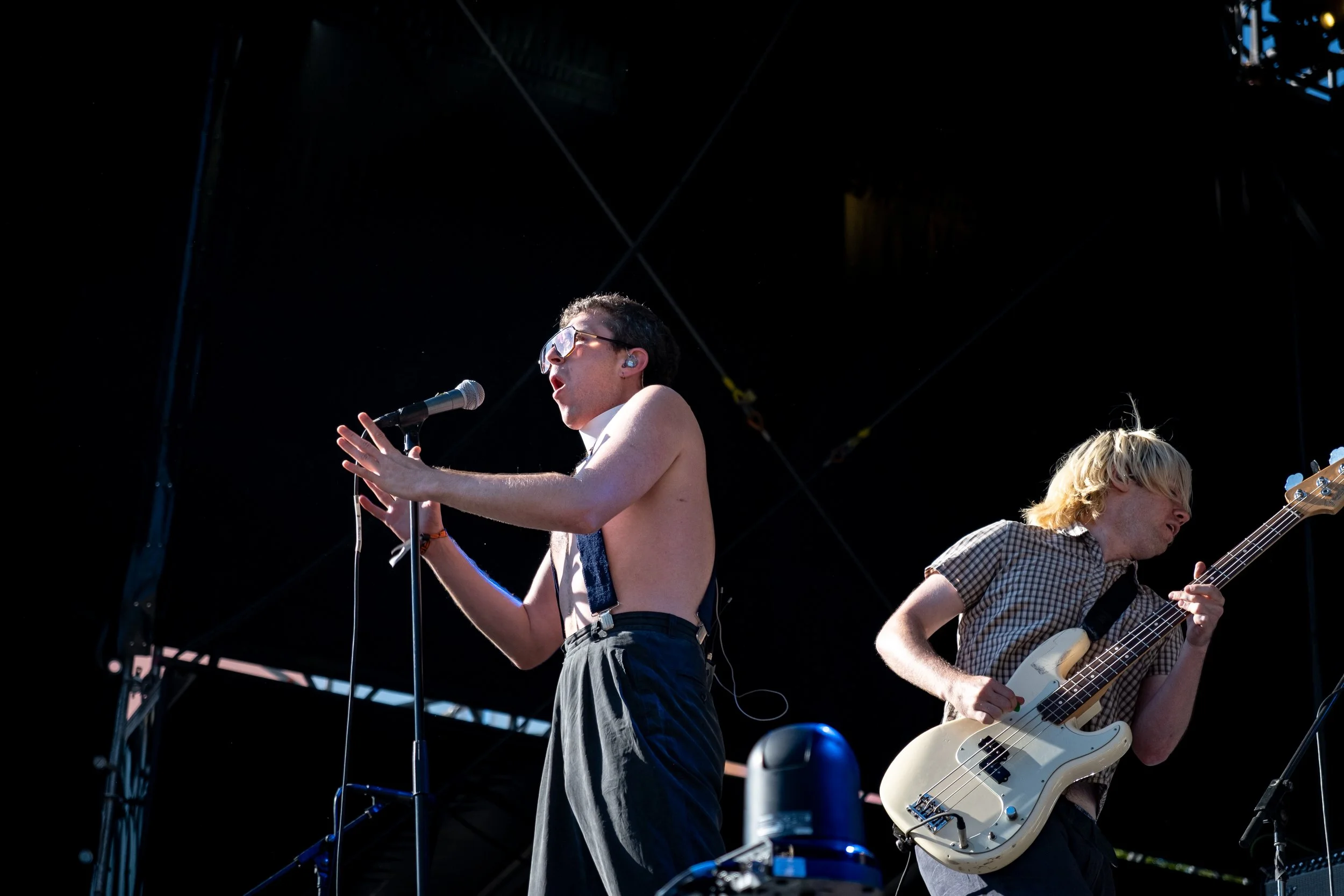 Two band members perform on a stage: one singing into a microphone and the other playing an electric bass guitar, with a dark stage background and visible stage equipment.