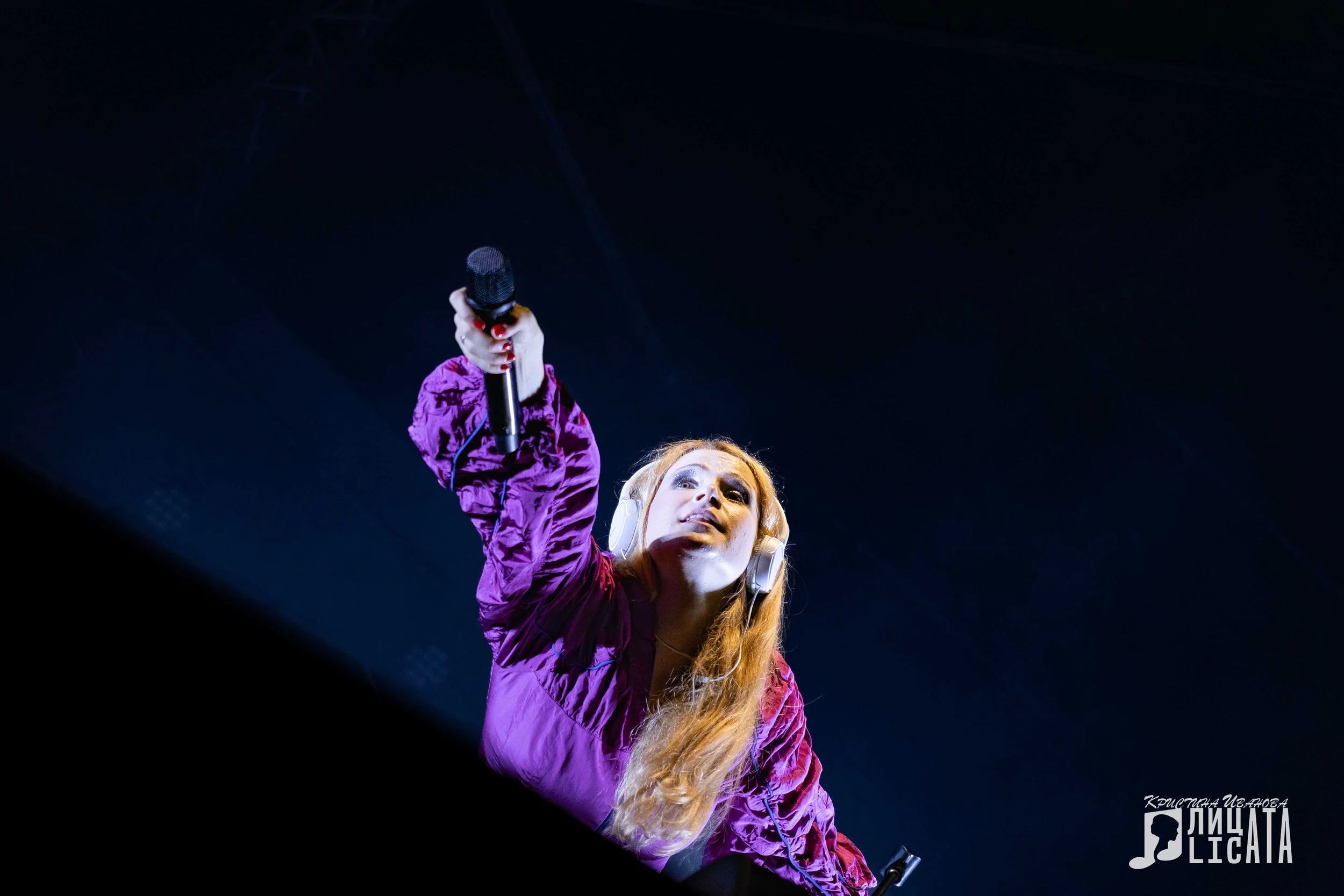 A female DJ with long blonde hair wearing headphones and a purple jacket, holding a microphone and performing on stage under blue lighting.