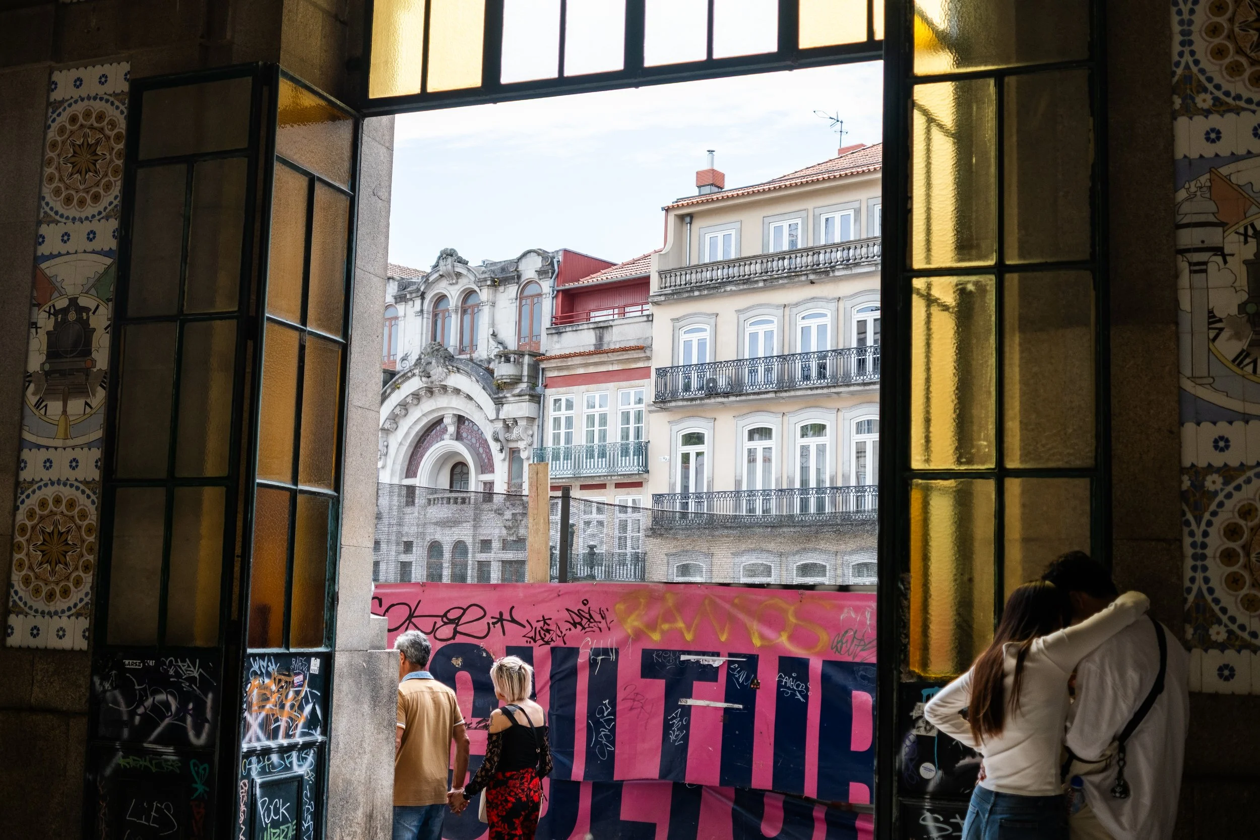 View through an open archway showing a city street with old buildings, a pink graffiti-covered barrier, and two couples standing close together, embracing or talking.