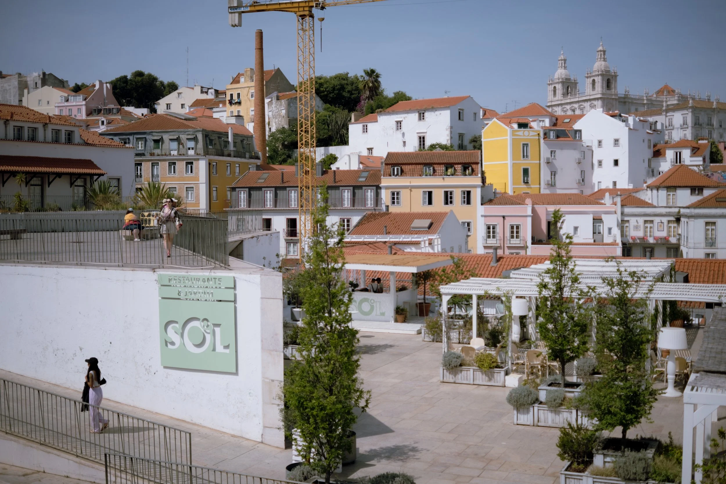 Colorful hillside neighborhood with pastel-colored buildings, red-tile roofs, and a church with twin towers in the background. In the foreground, a white wall with a restaurant sign and outdoor seating area with trees.