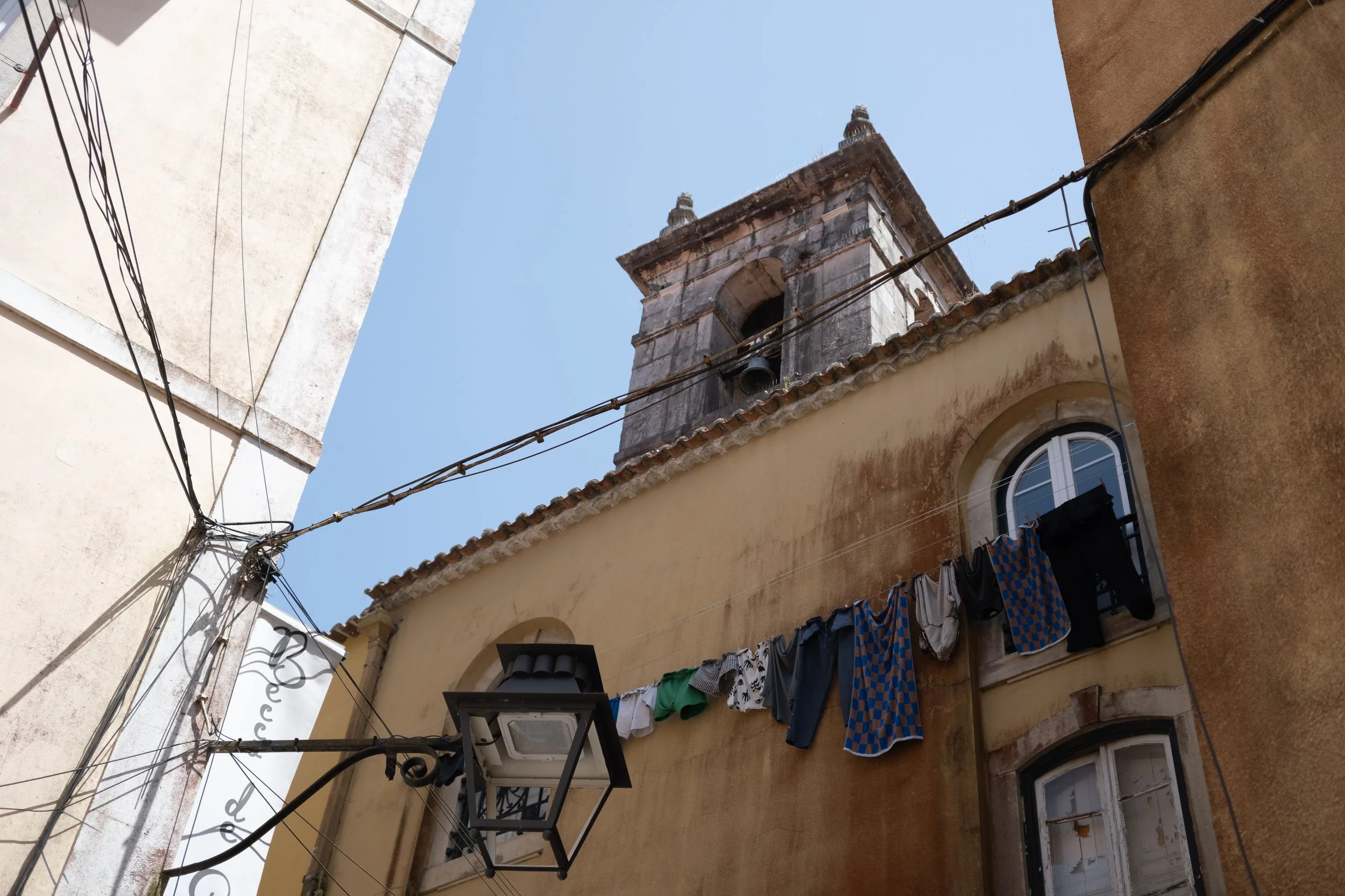 View of an old church tower against a clear blue sky, with laundry hanging on a wire between surrounding buildings.