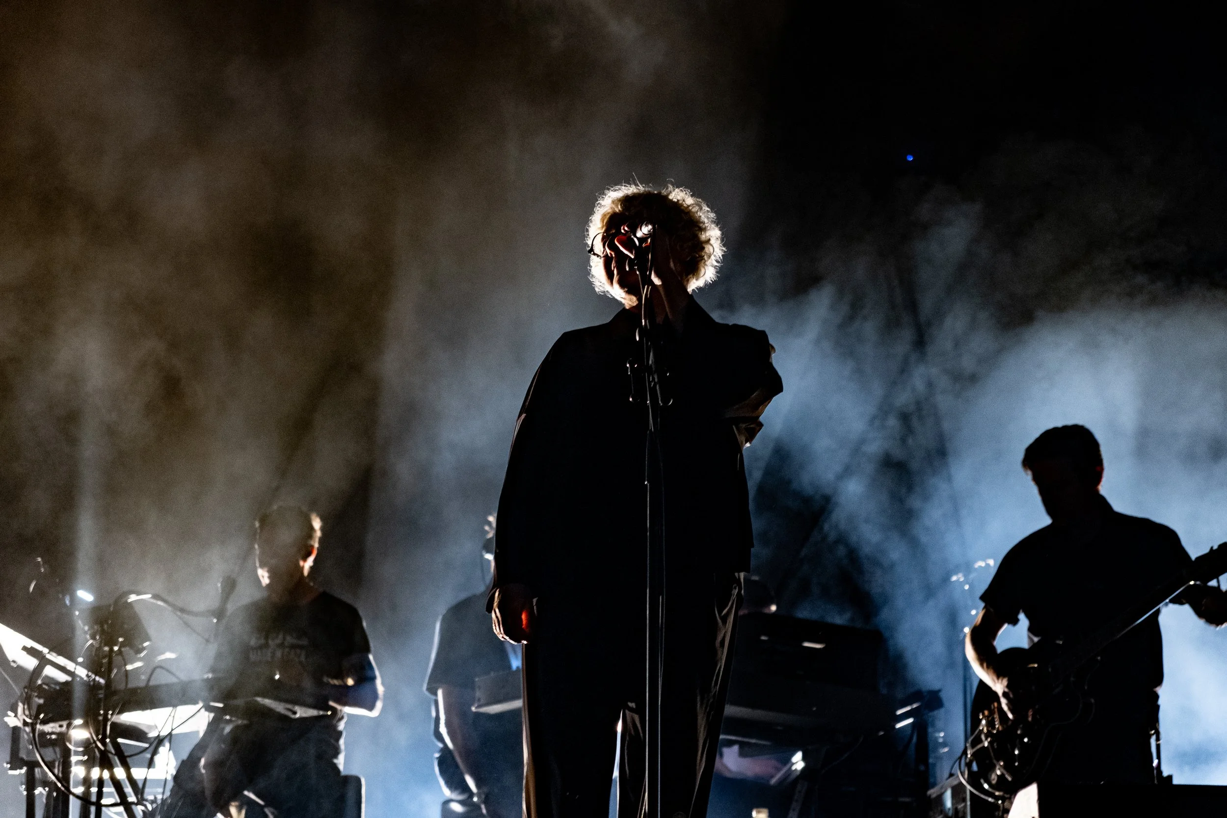 A silhouette of a person with curly hair singing into a microphone on stage, with band members playing instruments in the background, illuminated by stage lights and surrounded by smoke.