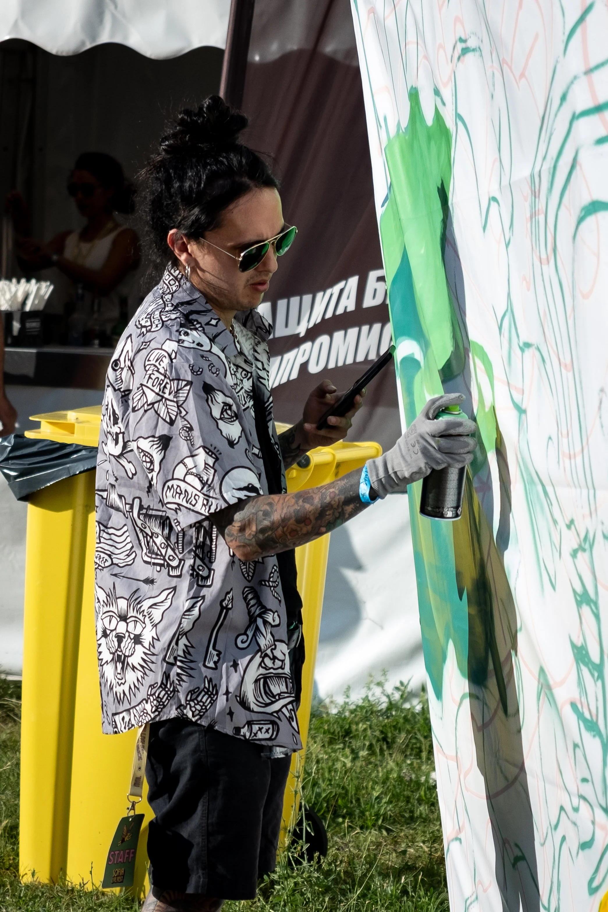 A man with dark hair, tattoos, sunglasses, and a patterned shirt is spray painting on a large canvas with green and orange colors. He holds a spray paint can in his right hand and a phone in his left. Behind him, there are yellow trash bins and a ten