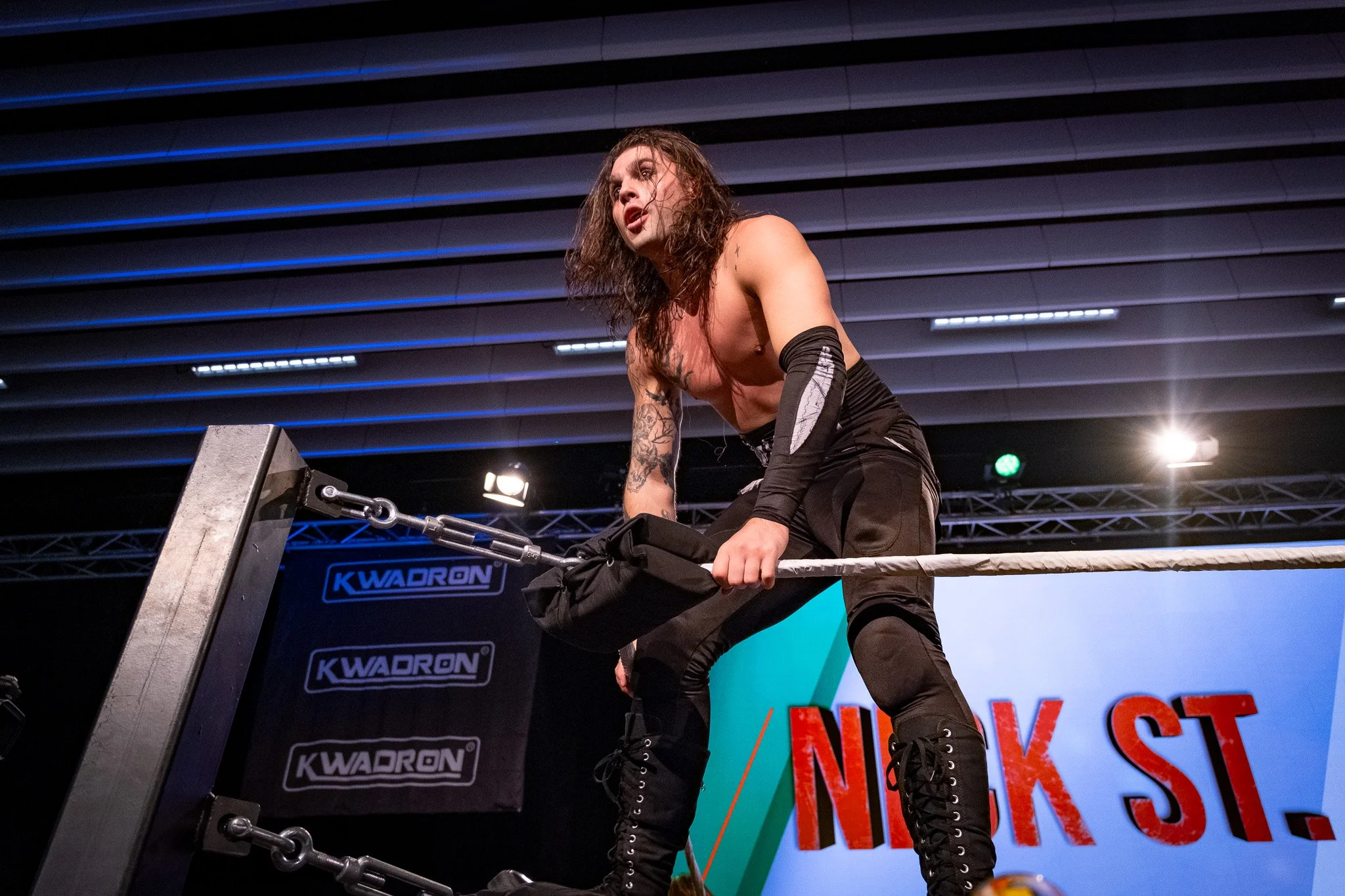 A female wrestler with long curly hair, injured arm with a bandage, and tattoos, wearing black pants and boots, leaning on the middle rope of a wrestling ring during a match, with bright stage lights and a large screen displaying text in the backgrou