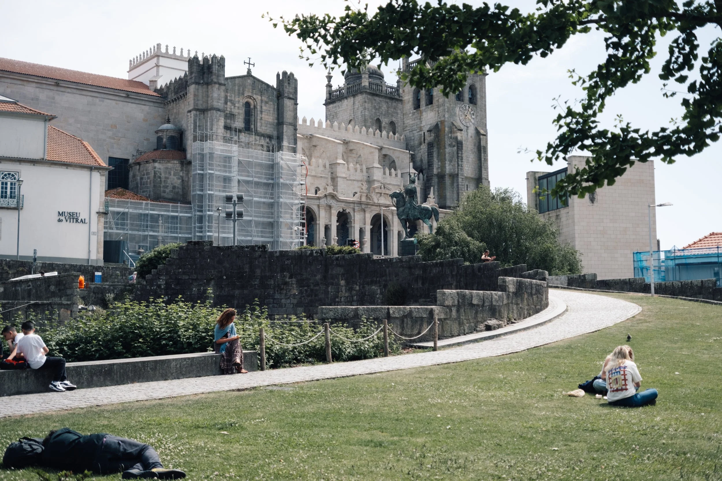 People sitting and relaxing on grass and benches in front of a historic stone building with scaffolding and an equestrian statue.