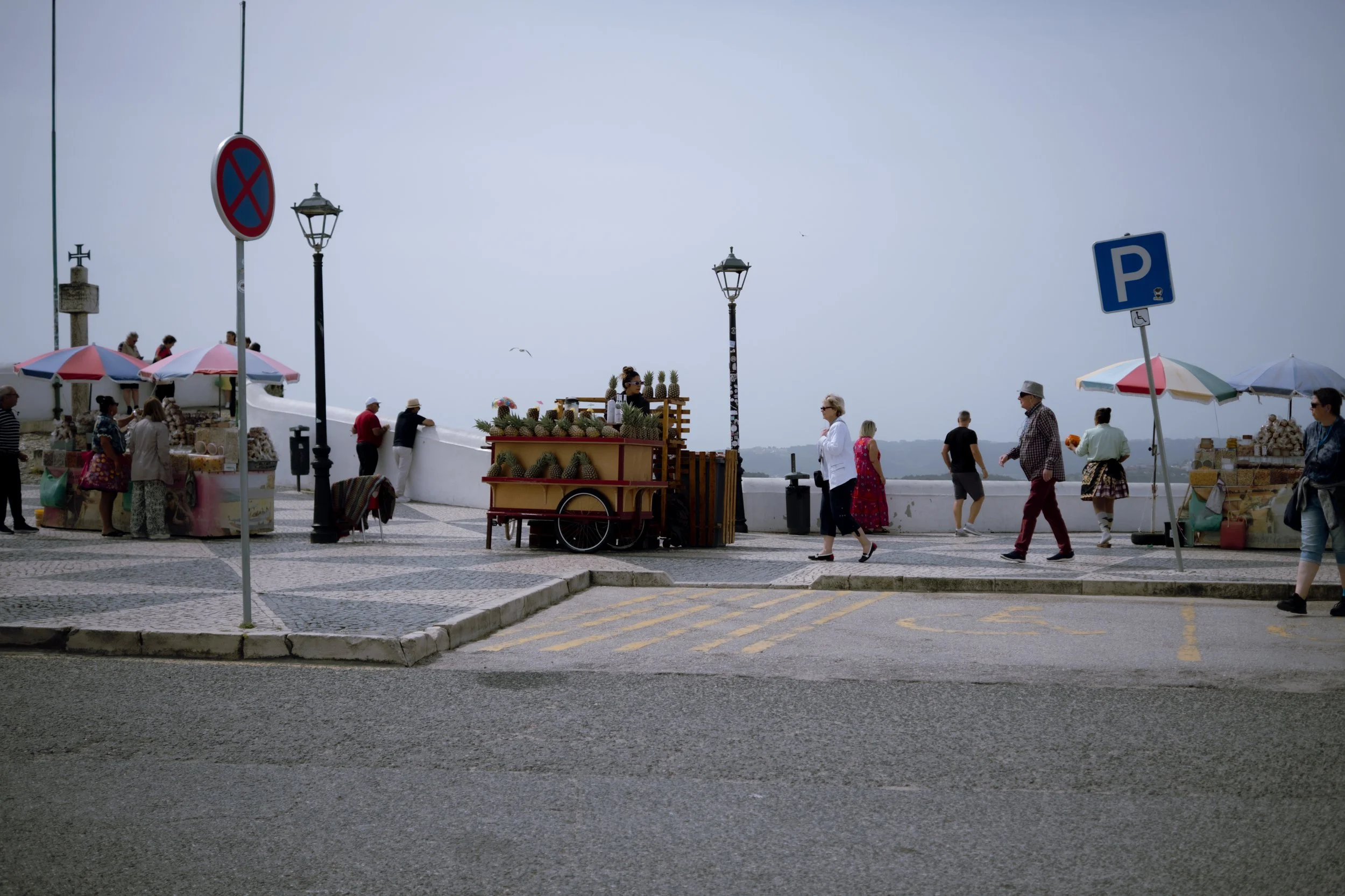 People walking and shopping at an outdoor market along a seaside promenade with street lamps and umbrellas, overcast sky in the background.