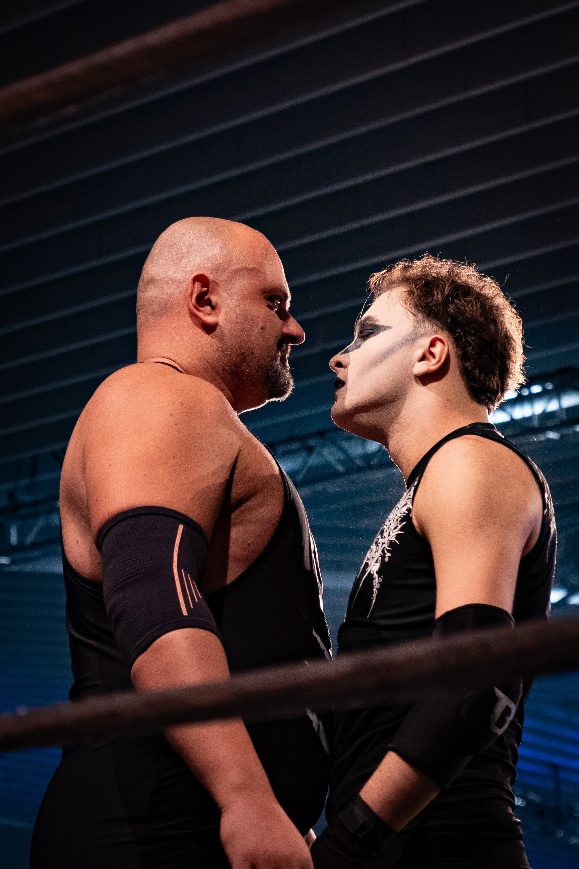 Two wrestlers face each other in a wrestling ring, one with a shaved head and the other with face paint and short hair.
