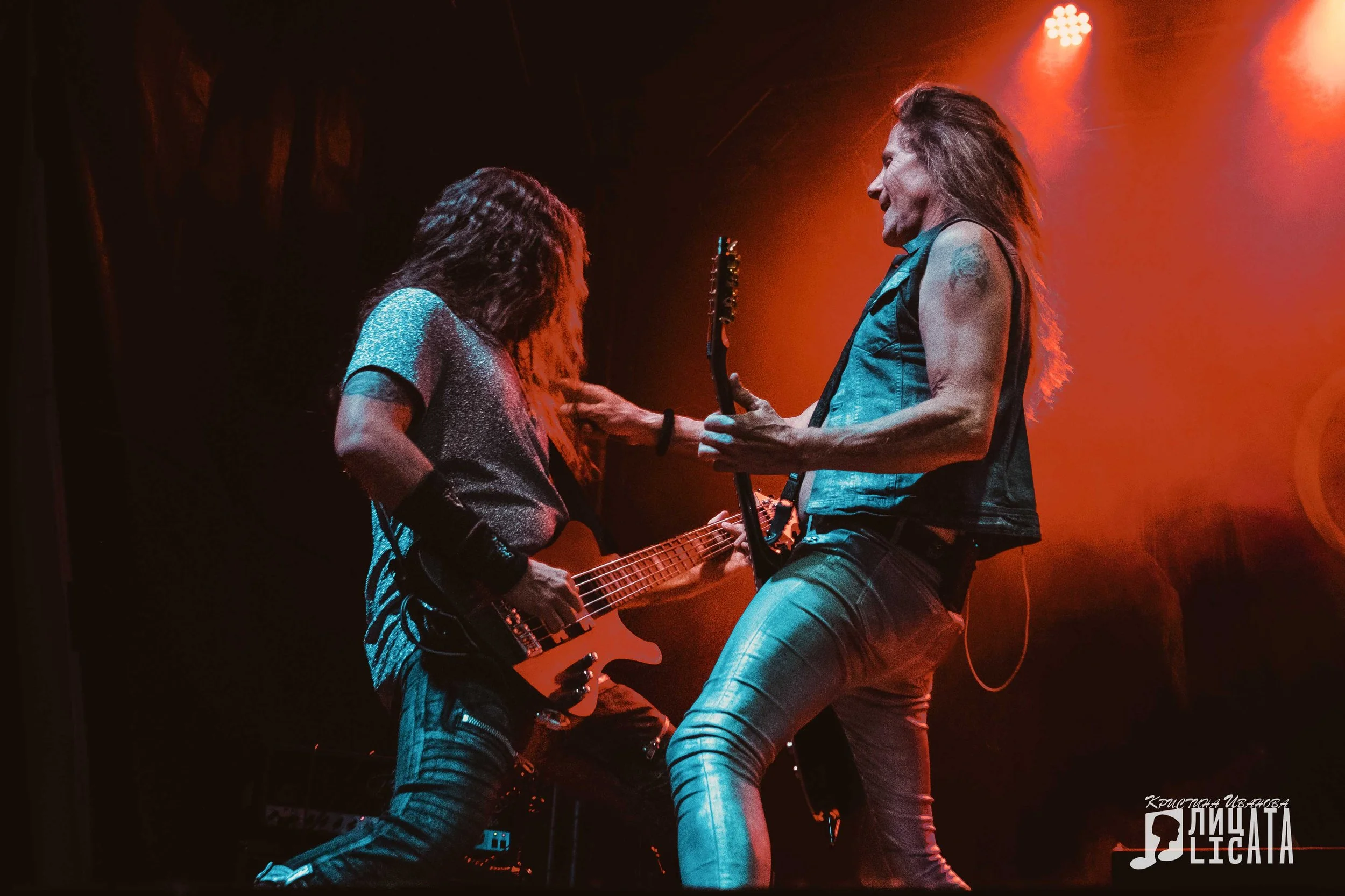 Two rock musicians performing on stage, playing guitars under red stage lighting.