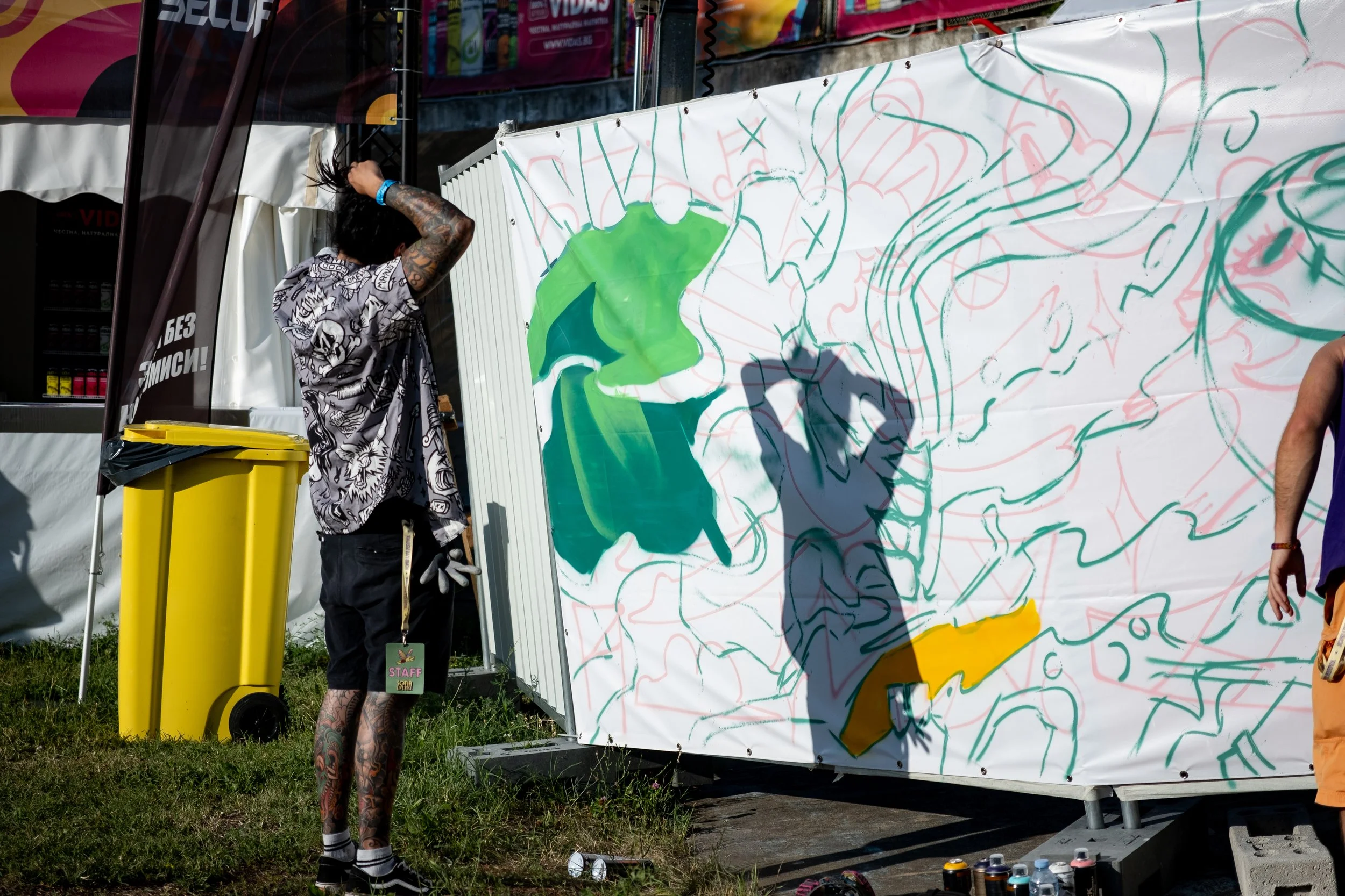 An artist with tattoos and long dark hair, wearing a black and white patterned shirt, is standing outdoors, working on a large abstract graffiti mural with green, dark blue, yellow, and red spray paint on a white canvas. There is a yellow trash bin n