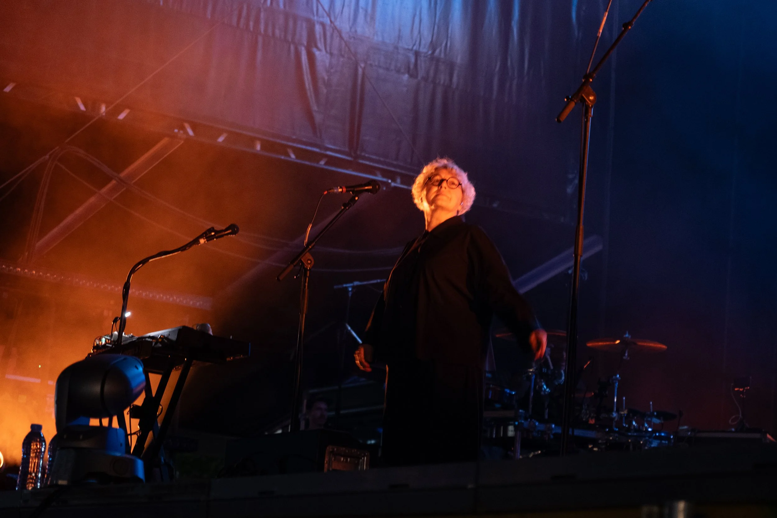 Elizabeth Fraser with Massive Attack at Sofia Live Festival. A person with white hair and glasses standing on a stage with musical equipment, illuminated by colorful stage lights, facing slightly to the right.
