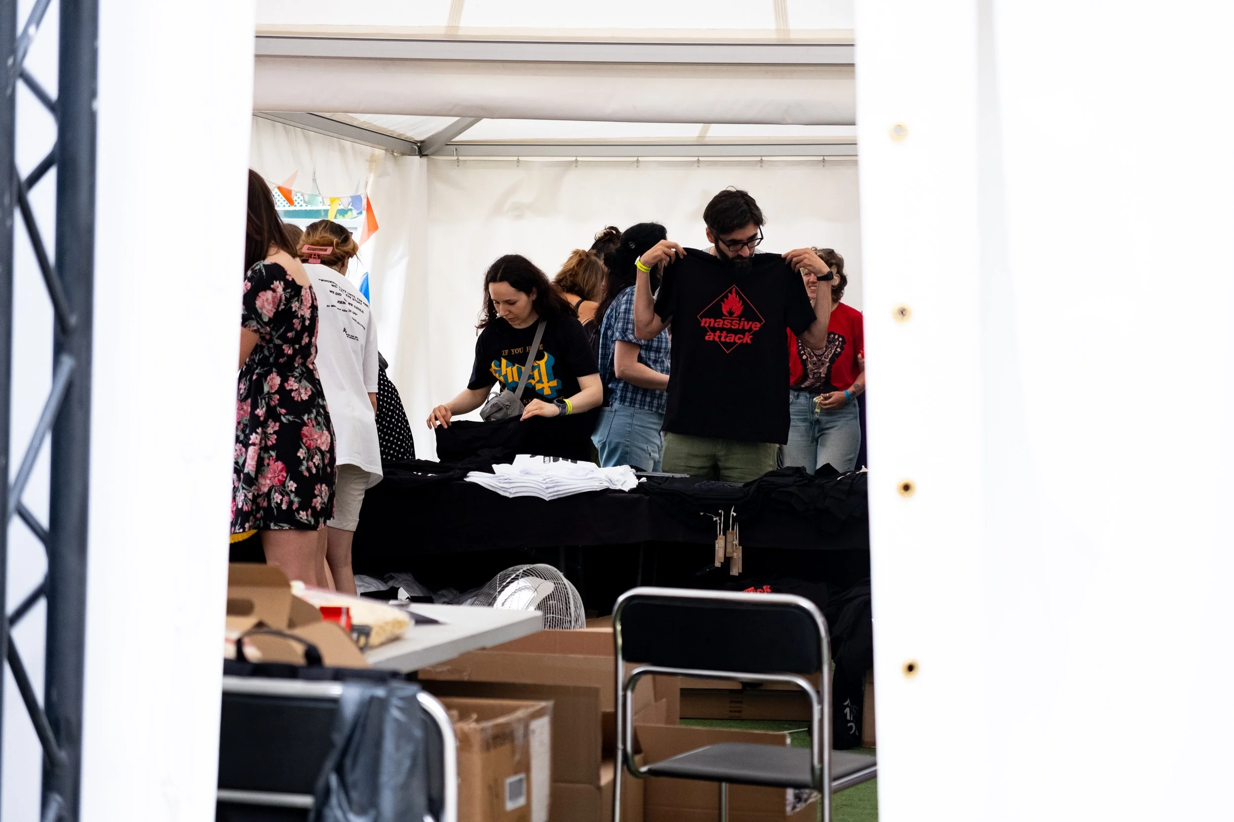 Group of people inside a tent, browsing and organizing shirts on a table, with cardboard boxes and chairs in the foreground.