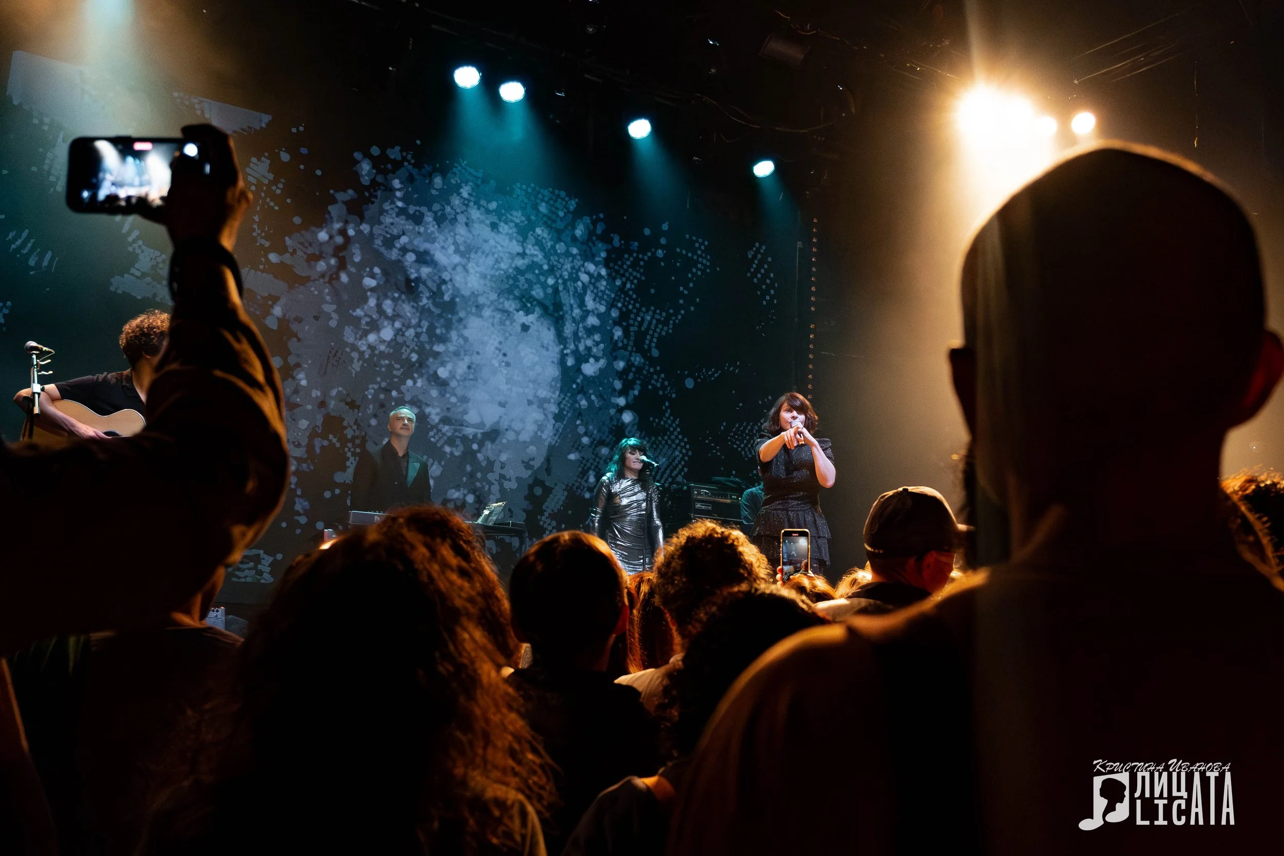 A live concert with a female singer performing on stage, accompanied by musicians, under colorful stage lights, with an audience watching and taking photos.