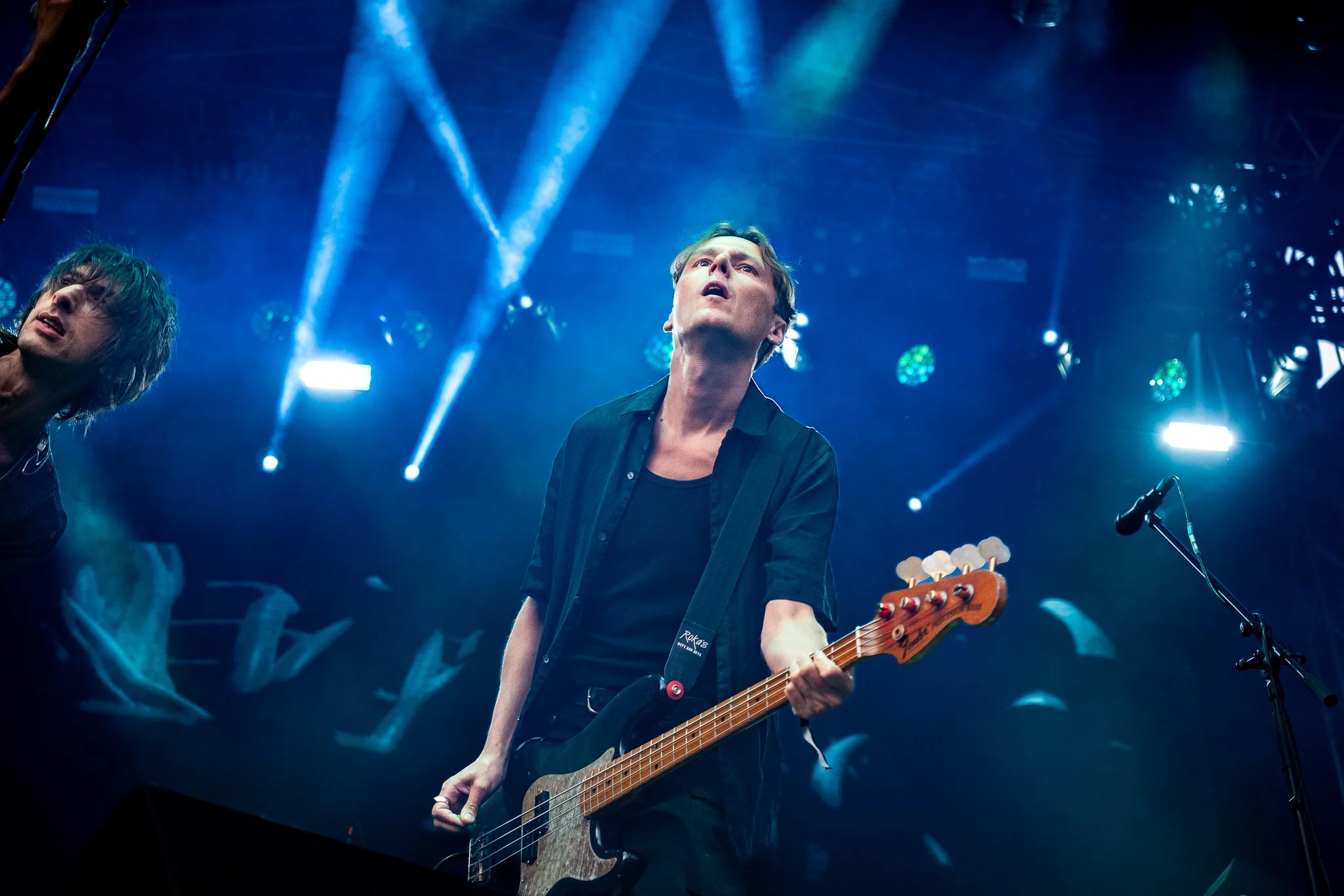 The Horrors band at Sofia Live Festiva. A male musician playing an electric bass guitar on stage during a live performance, with blue stage lighting and a microphone to his right.