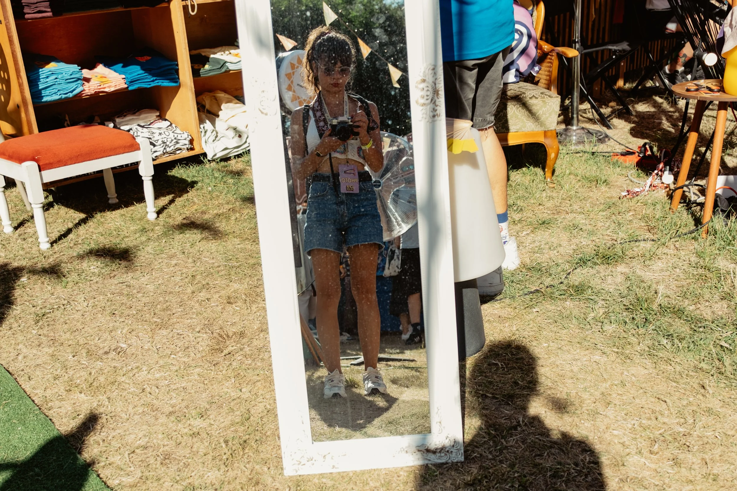 A woman with curly hair wearing sunglasses, a tank top, and denim shorts taking a photo in a tall, ornate full-length mirror outdoors at a flea market or yard sale.