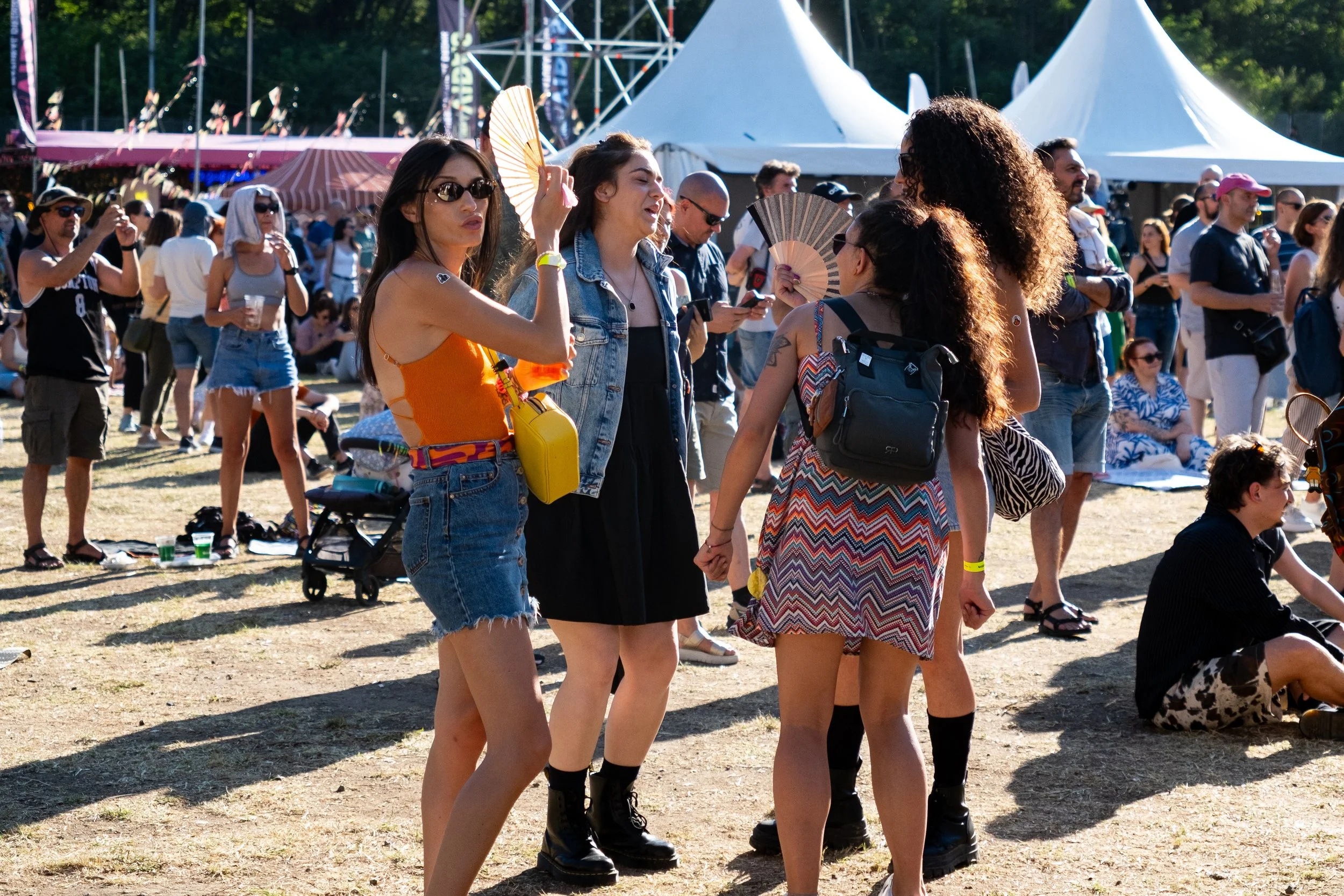Group of people attending an outdoor music festival or fair, standing on grass with white tents and a stage in the background, some talking, taking photos, and holding fans.