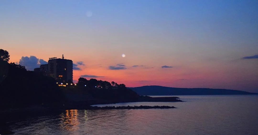 Sunset over a body of water with silhouettes of buildings and trees on the shoreline, pink and purple sky with some clouds and the moon visible.