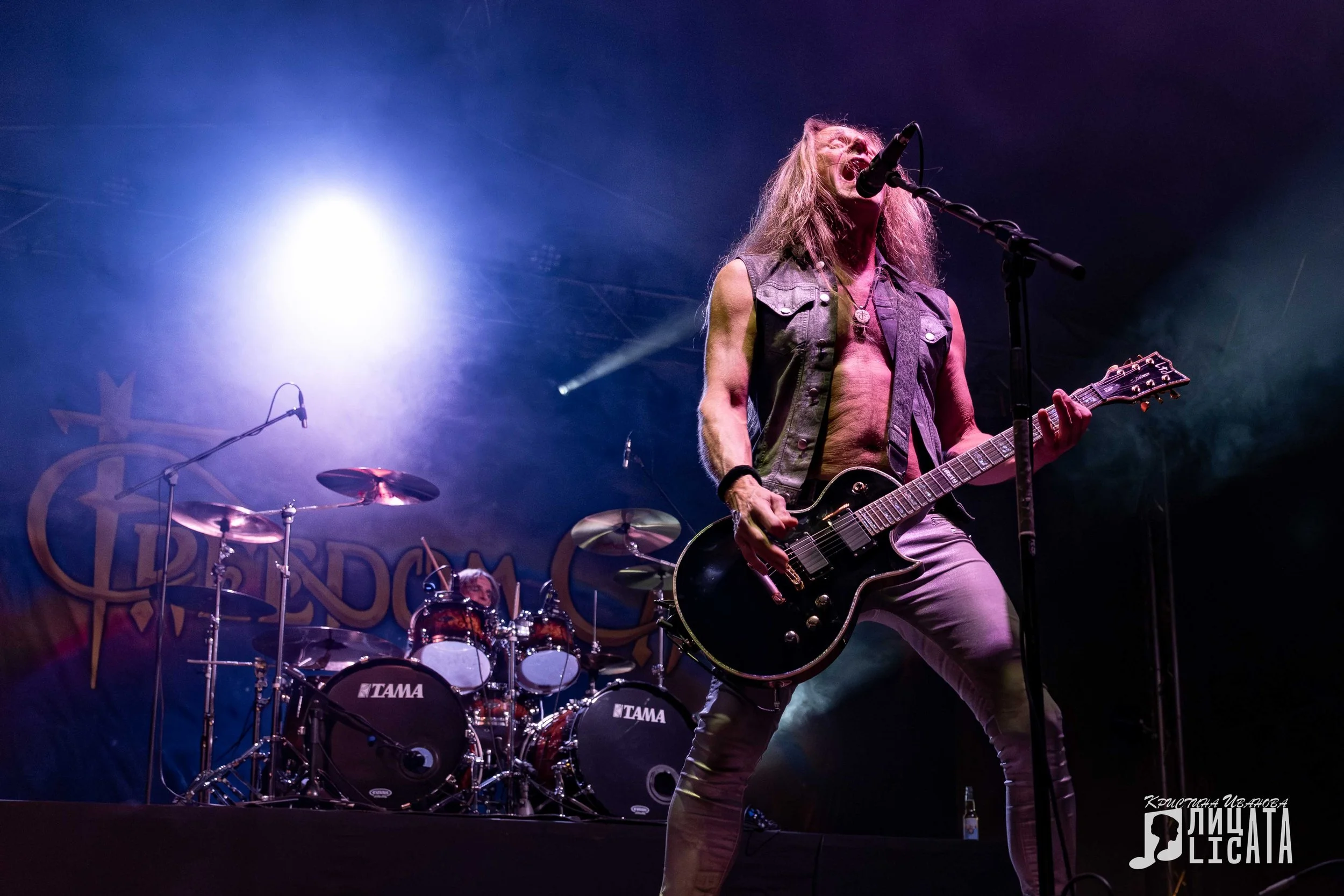 Male rock musician with long hair, sleeveless vest, and guitar performing on stage with drummer in the background and stage lighting.