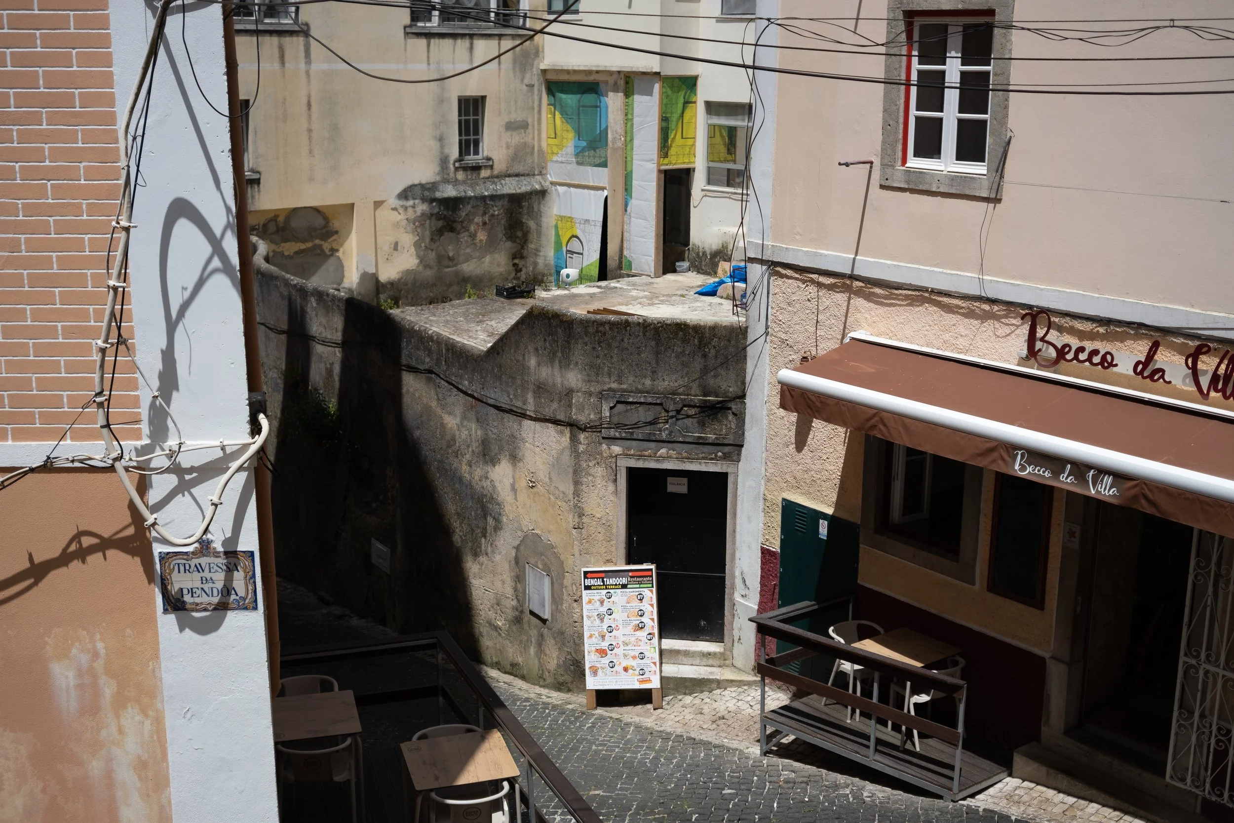 View of a narrow street corner in an urban area with a restaurant on the right, featuring a brown awning reading 'Becco da Villa', and outdoor seating. There's a menu board outside, a yellow wall with a door and window, and overhead power lines.