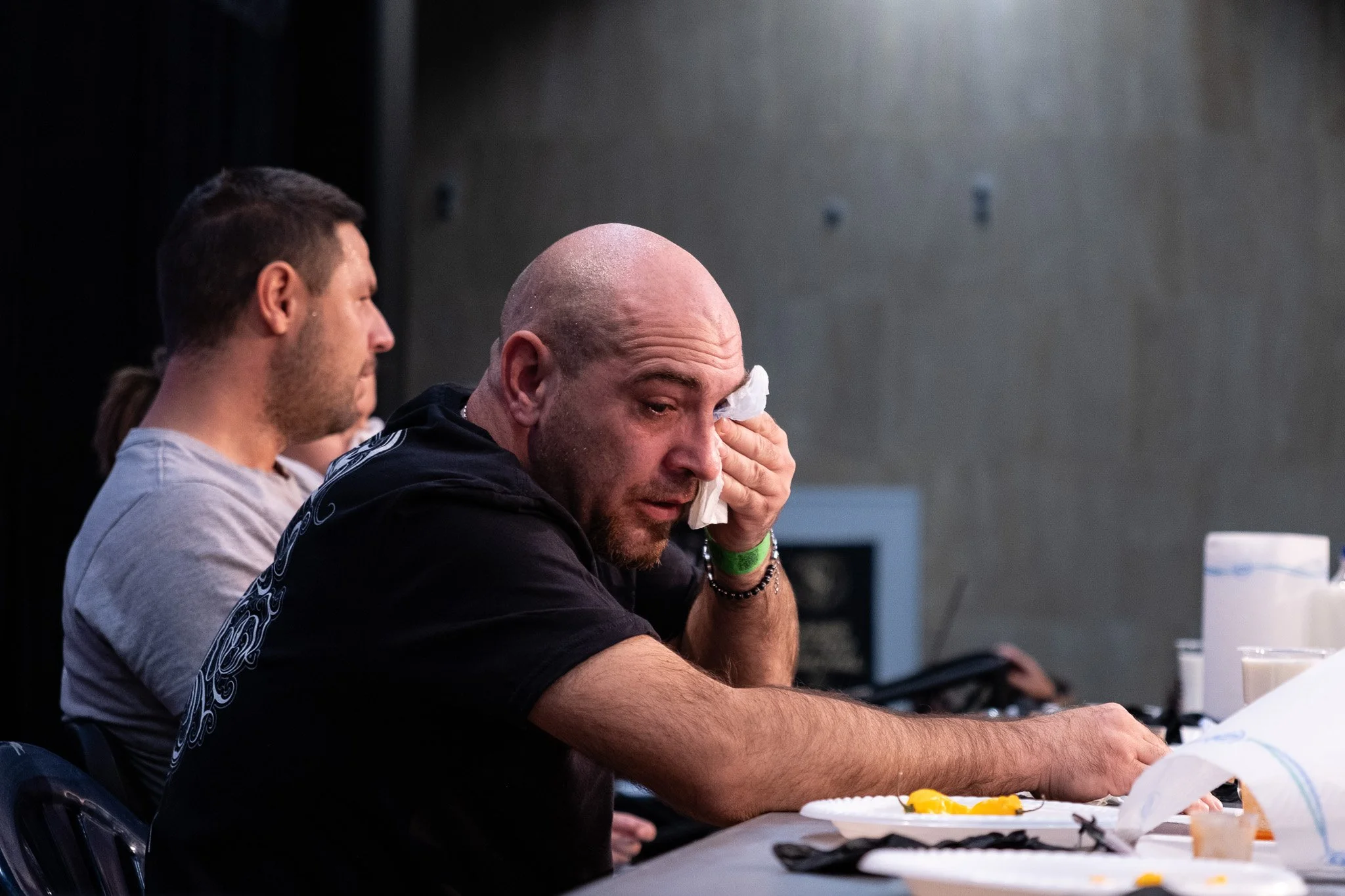 A man with a shaved head is cleaning his right eye with a tissue, sitting at a table with food and drinks, while another man with long hair tied back watches. The setting appears to be a casual indoor event or workshop.