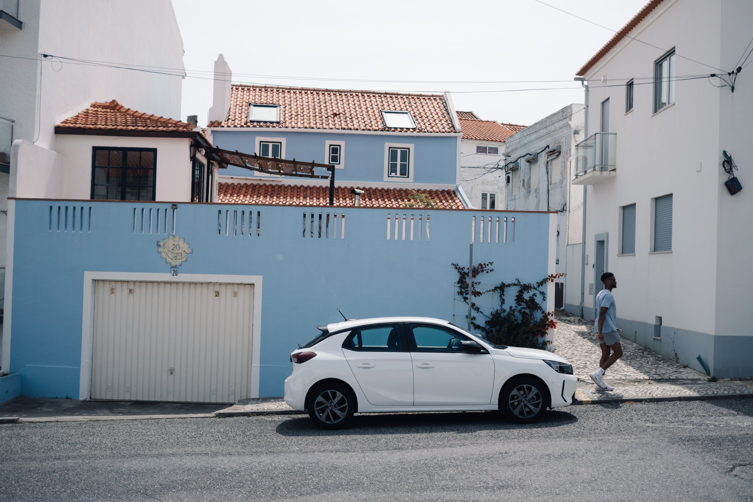 A white car parked in front of a blue building with a garage door. A man in a white shirt and gray shorts is walking past the car on a cobblestone sidewalk. The building has a red-tiled roof, with other buildings in the background, and a vine growing