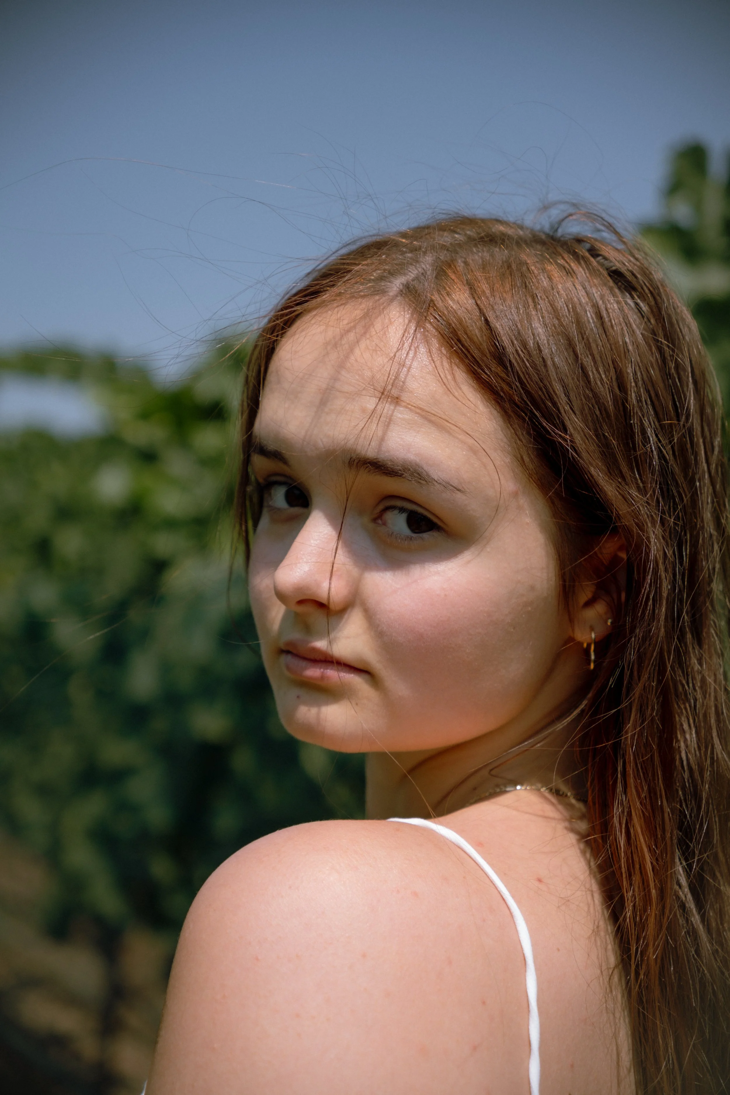 A young woman with light skin and reddish-brown hair outdoors on a sunny day, looking over her shoulder at the camera with a slight smile, with green foliage and a blue sky in the background.