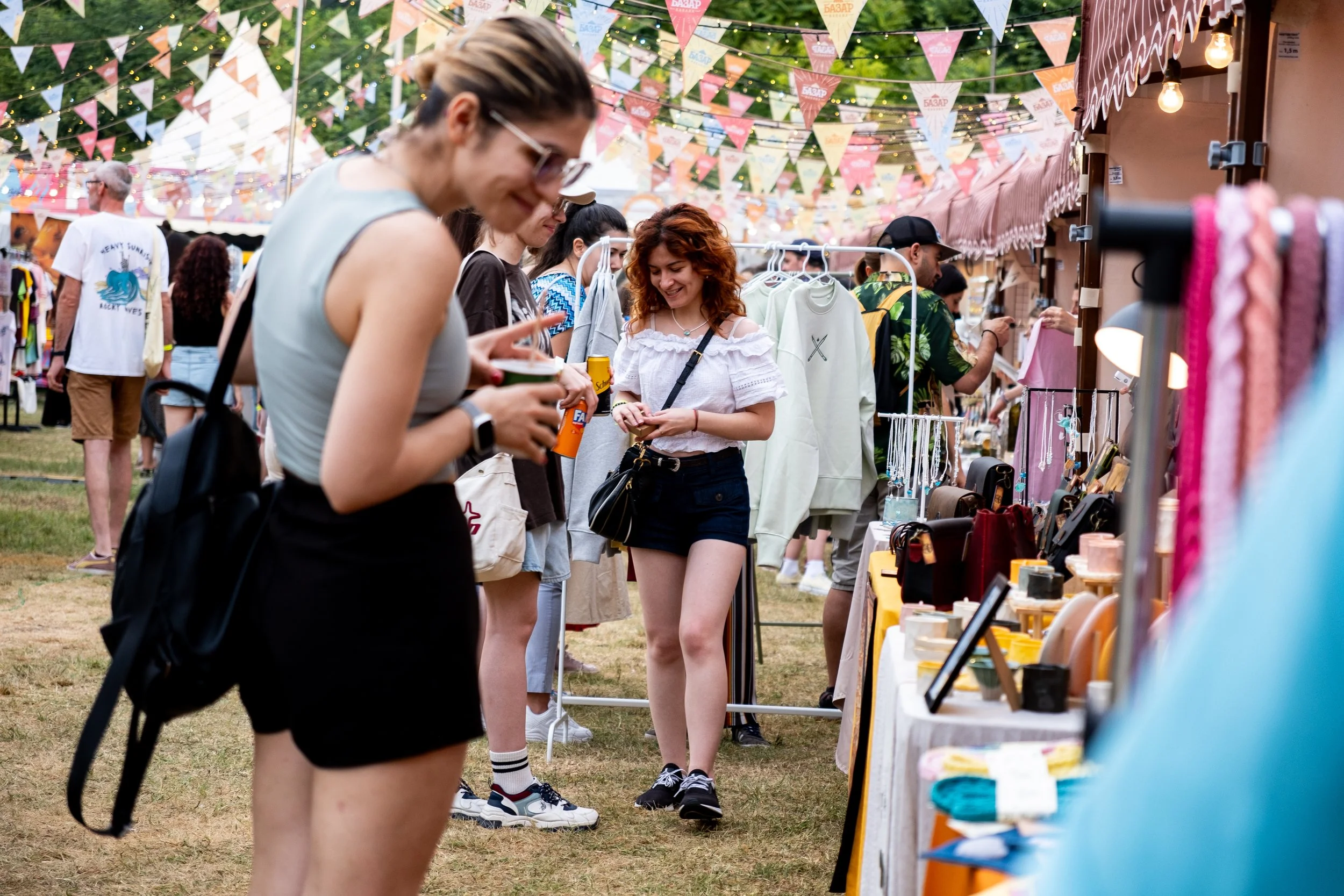 People shopping at an outdoor market decorated with colorful banners and string lights, browsing clothing and jewelry stalls.