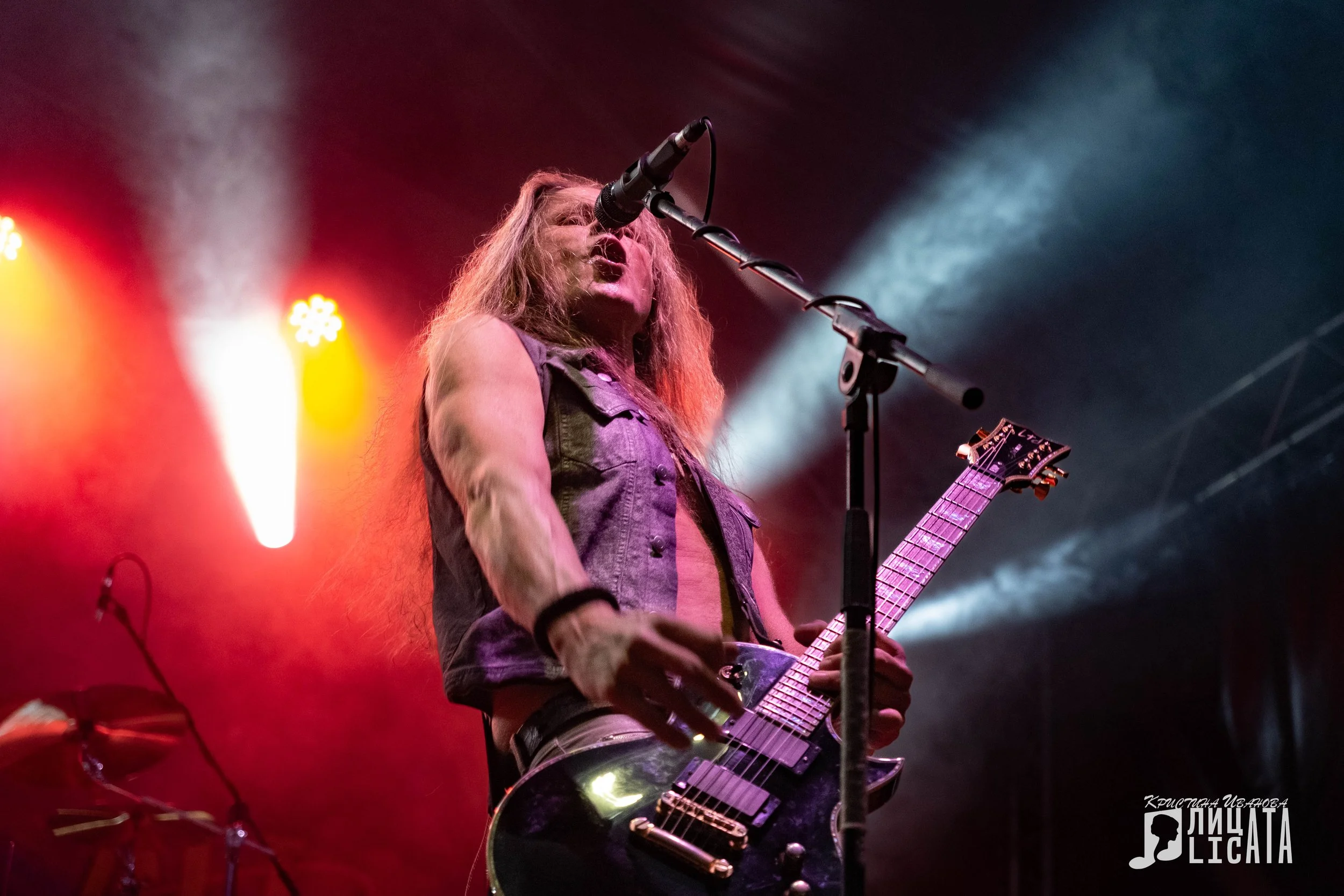 A male guitarist and singer performing on stage, playing a black electric guitar and singing into a microphone, with colorful stage lights in the background.