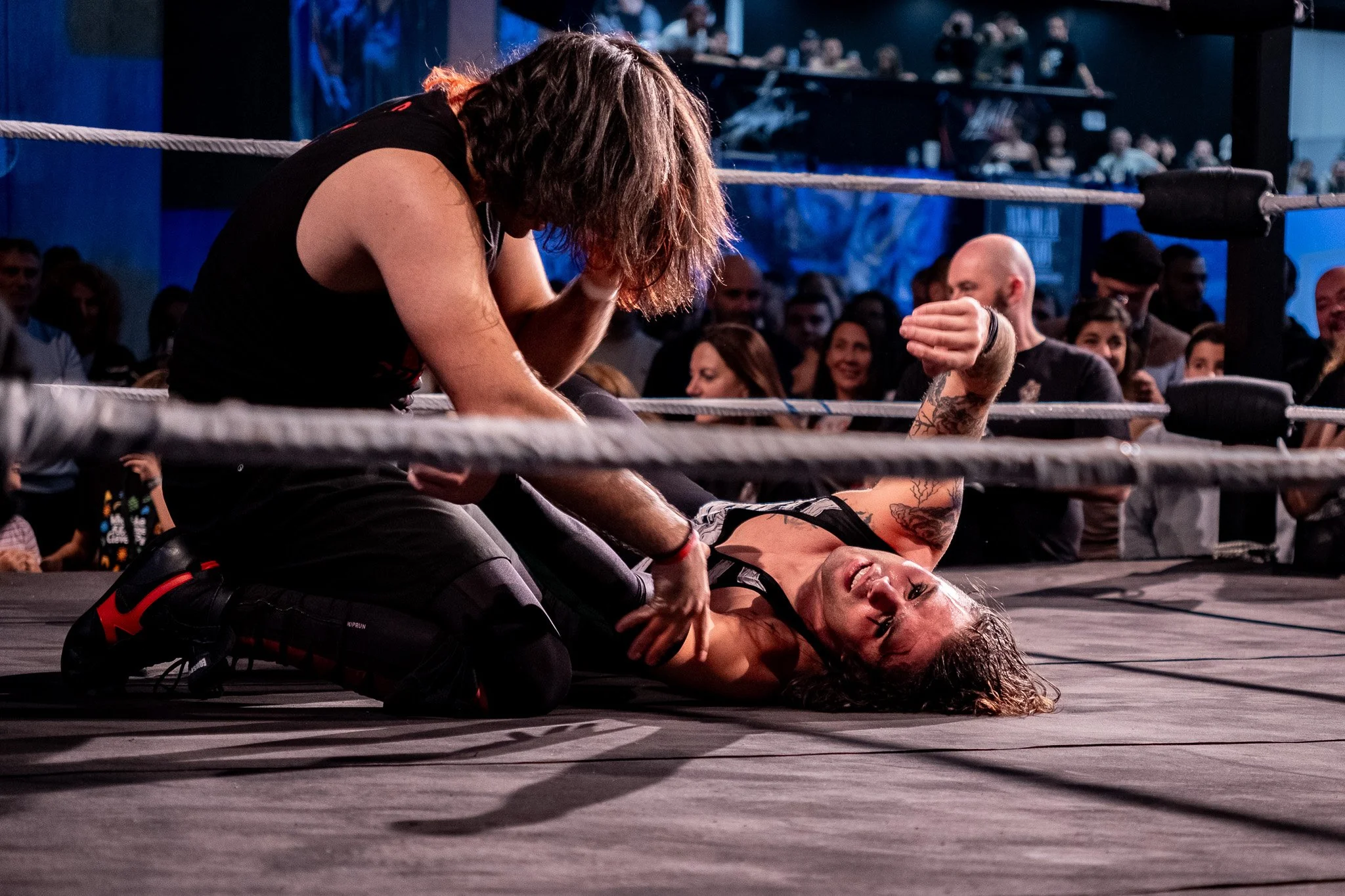 Wrestling match in progress with a female wrestler on the mat and a male wrestler kneeling beside her, surrounded by a crowd of spectators.