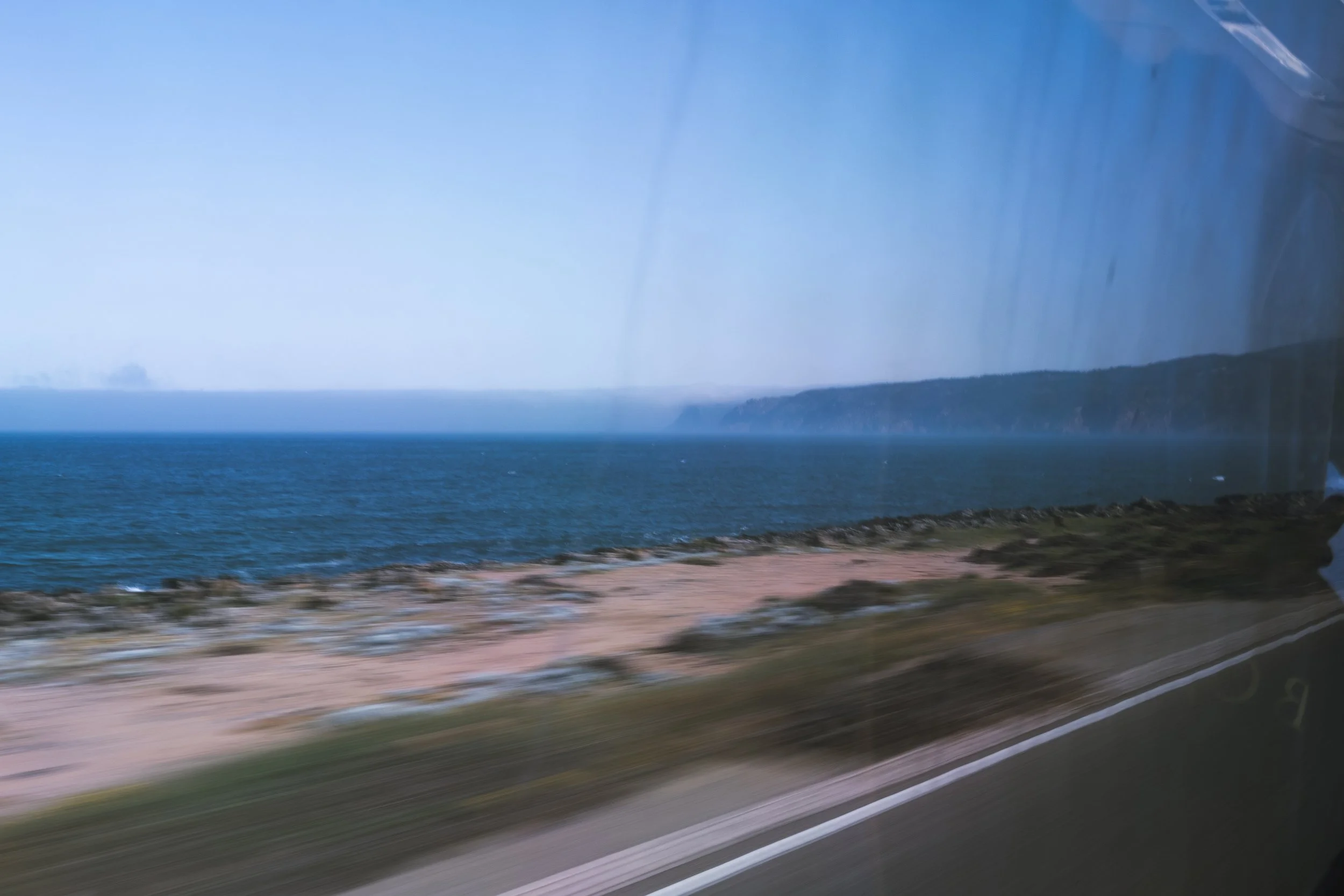 A view of the ocean coastline with water and greenery, seen through a car window with motion blur.