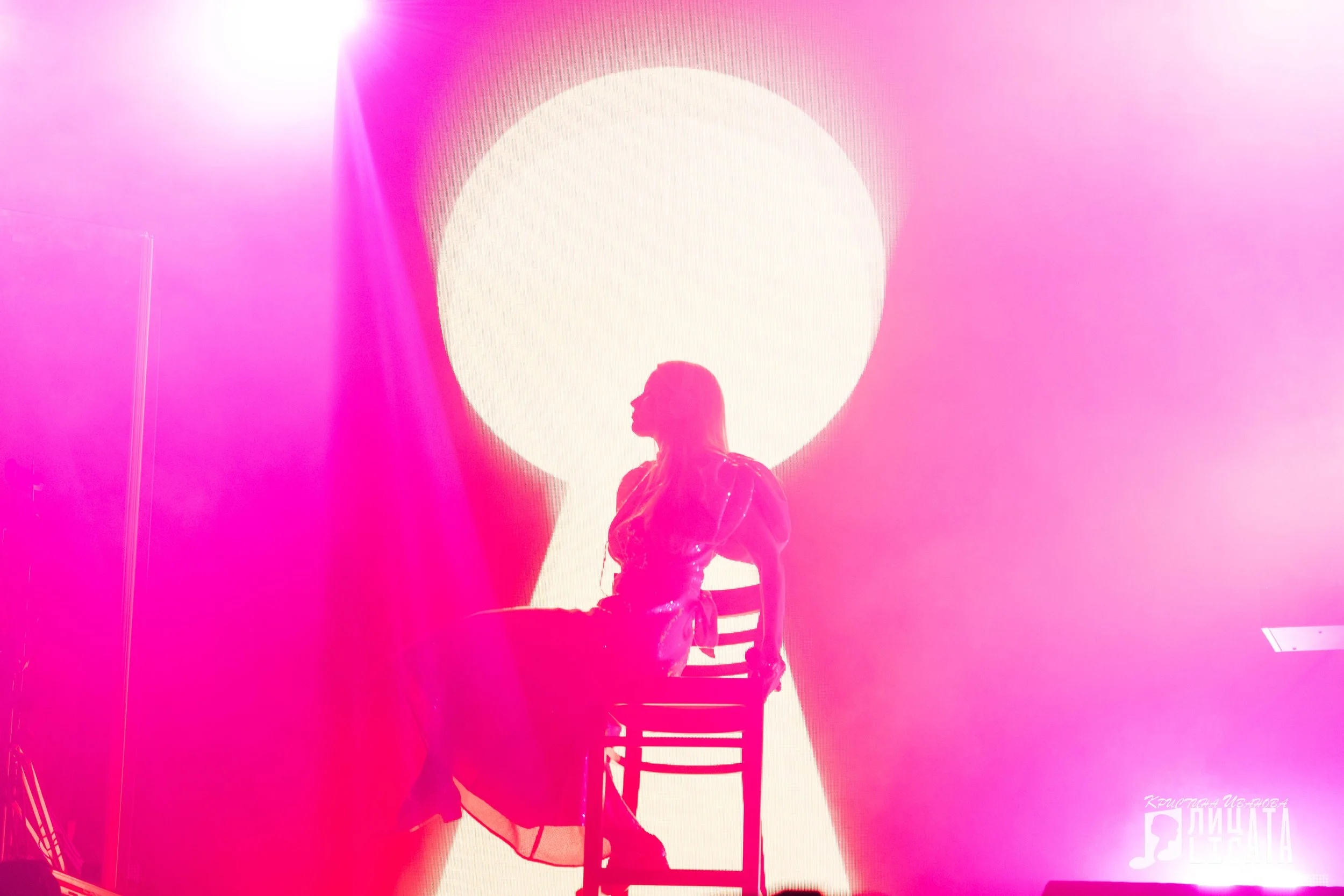 A woman sitting on a chair on stage, backlit by a large bright circle resembling a keyhole, surrounded by pink and purple lights.